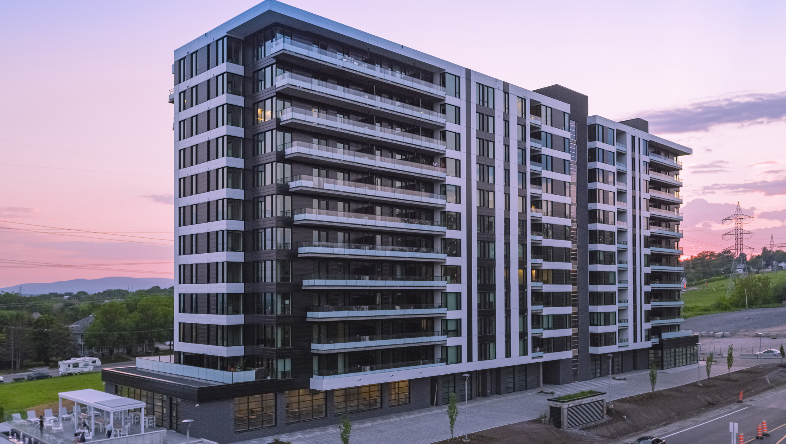 Une construction multirésidentielle moderne à plusieurs étages avec de nombreux balcons, de grandes fenêtres et un design épuré et contemporain se dresse au crépuscule. La scène est calme avec un ciel doux et rosé en arrière-plan et un paysage partiellement visible à gauche et à droite.