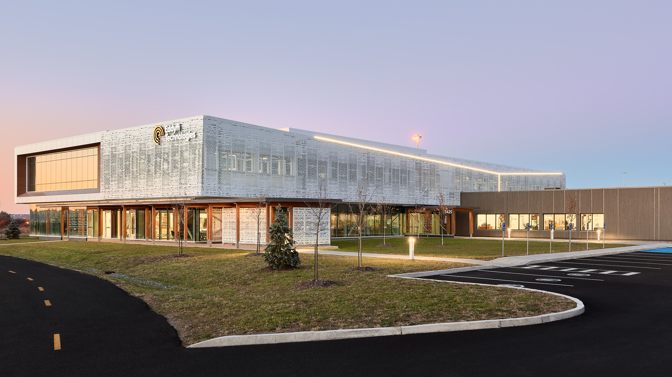 Un bâtiment moderne de deux étages avec une façade métallique épurée et de grandes fenêtres se détache sur un ciel crépusculaire. Le bâtiment est composé d'un mélange de verre et de métal perforé. Une allée incurvée et un aménagement paysager sont visibles au premier plan.