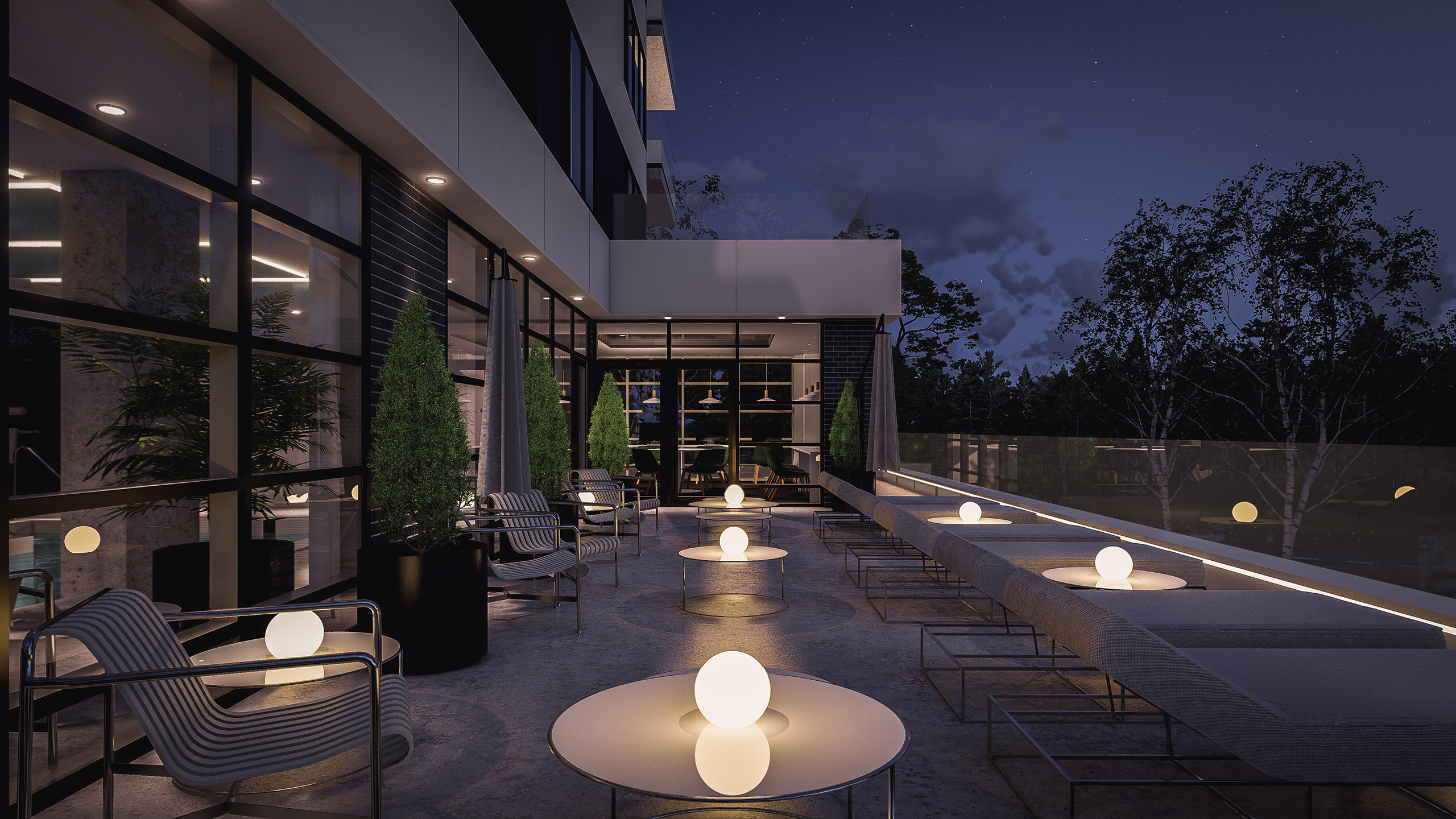 Une terrasse extérieure moderne, aménagée de nuit, avec un mobilier minimaliste, dont des tables rondes et des chaises rembourrées. De petites lumières sphériques sur les tables illuminent l'espace. De grandes plantes en pot et une balustrade en verre rehaussent le design épuré, caractéristique des constructions multirésidentielles sophistiquées, tandis que des arbres sont visibles en arrière-plan.