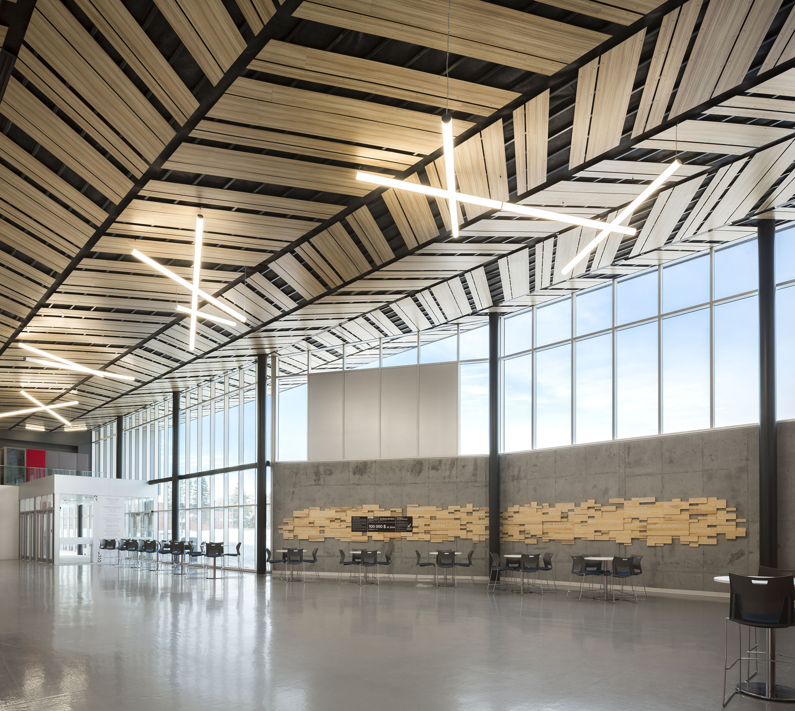 Un intérieur moderne et spacieux avec de hauts plafonds et de grandes fenêtres. Le plafond est doté de panneaux géométriques en bois uniques et de luminaires contemporains. Le mur de droite présente un mélange de carreaux de bois. L'espace est ouvert avec des tables et des chaises dispersées le long du mur.