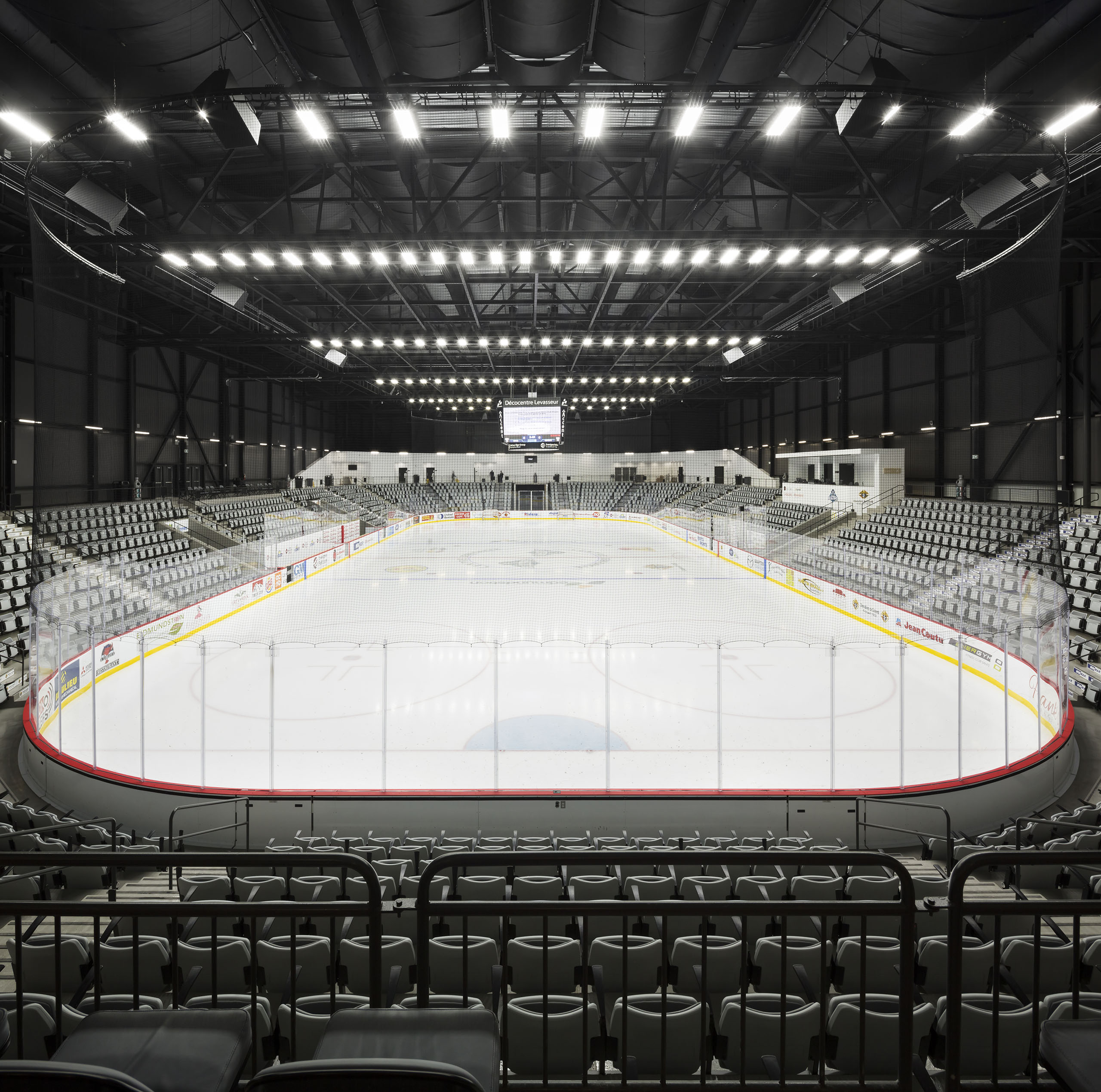 Une patinoire de hockey sur glace vide avec des lumières vives au plafond. La patinoire se trouve au centre, entourée de rangées de sièges gris, de barrières en verre et d'un tableau d'affichage suspendu au plafond. L'atmosphère est calme et immobile avant l'événement.