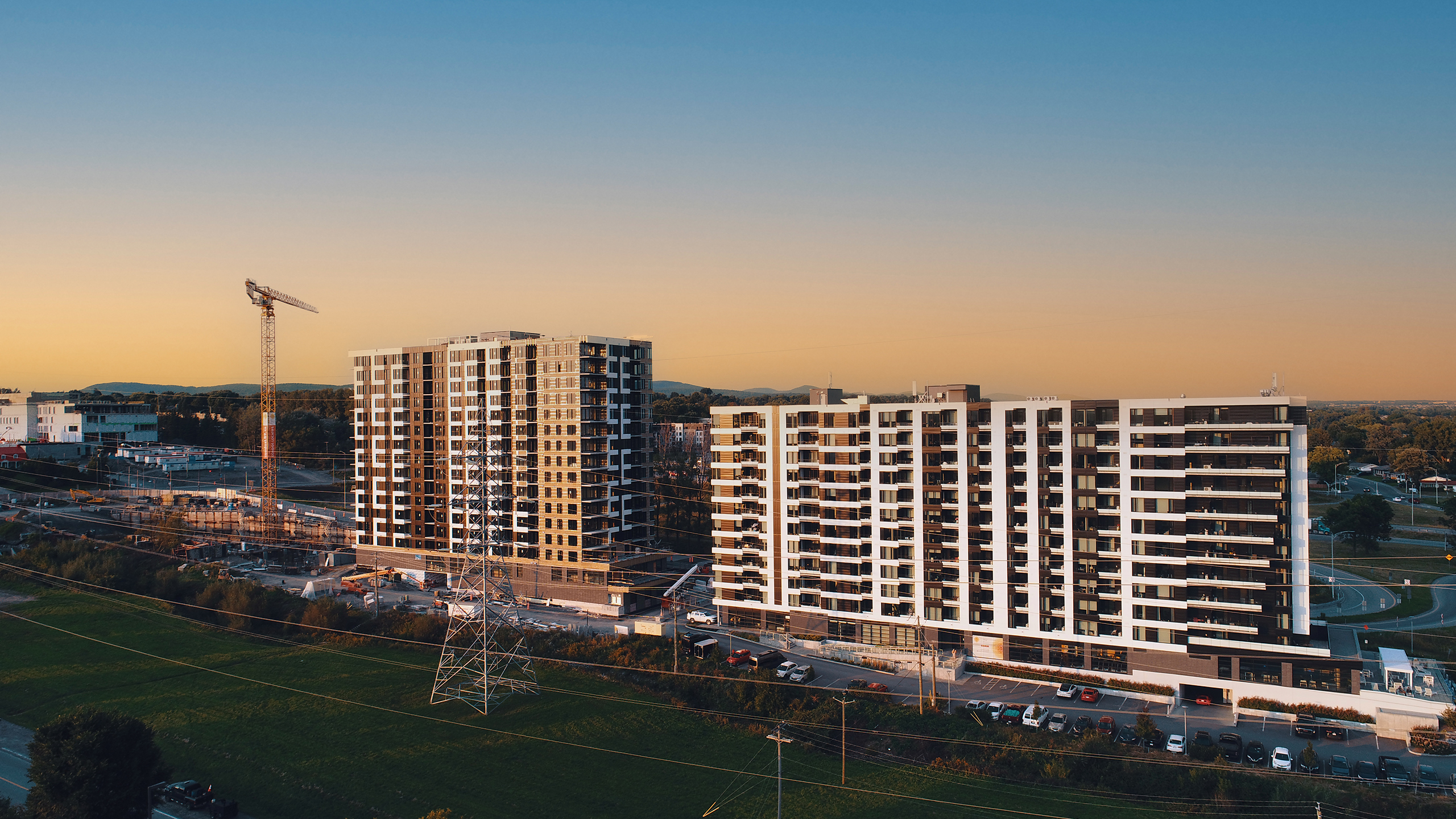 Vue aérienne de deux immeubles d'appartements modernes de grande hauteur en construction au coucher du soleil. Une grande grue et une structure partiellement construite sont visibles à gauche. Les environs comprennent des espaces verts, des routes, des voitures garées et des montagnes lointaines sous un ciel clair. Un instantané de la construction multirésidentielle en cours.