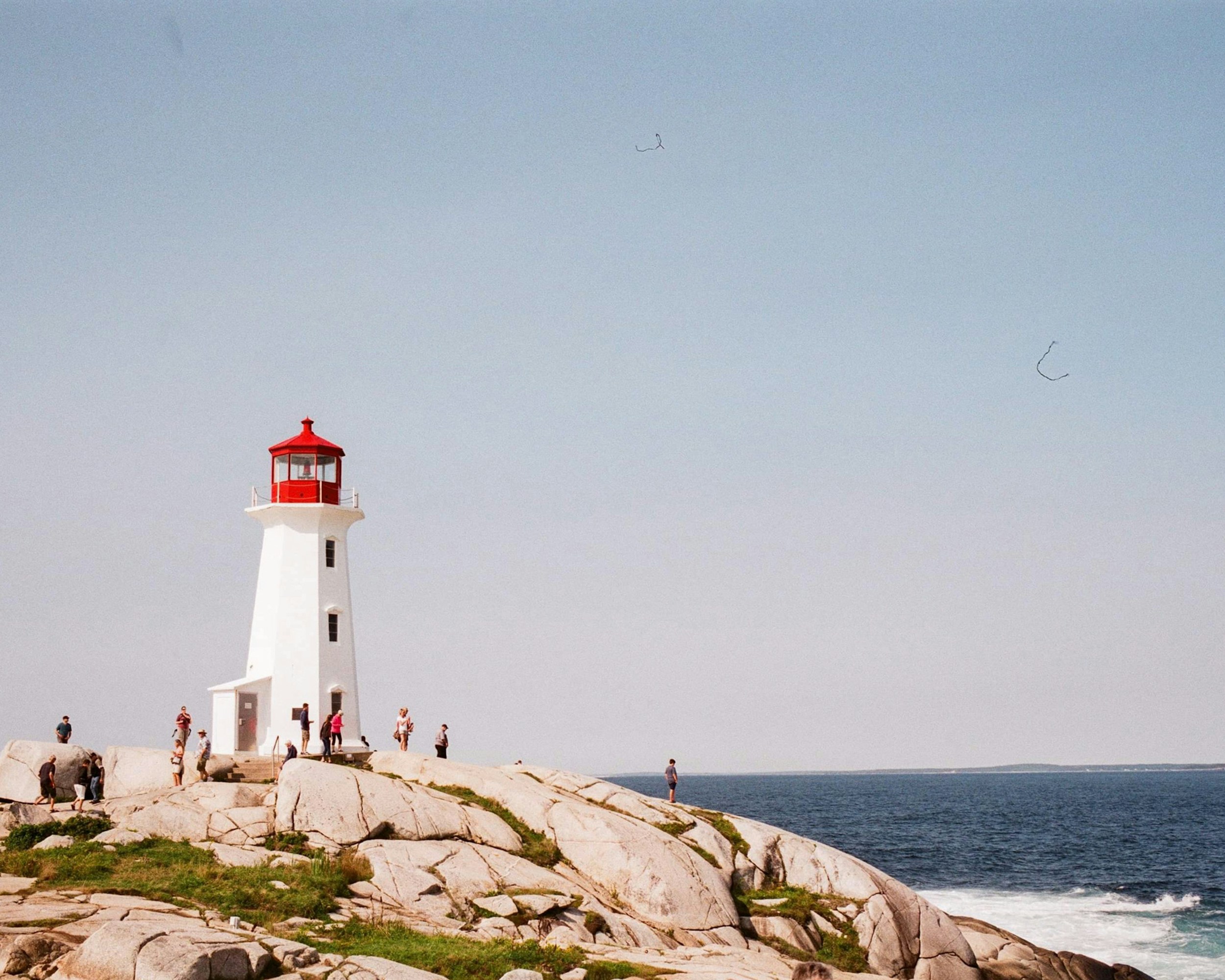 Un phare blanc au sommet rouge se dresse sur un rivage rocheux, sous un ciel bleu clair. Plusieurs personnes sont dispersées autour des rochers, certaines marchant, d'autres debout, avec l'océan visible en arrière-plan.
