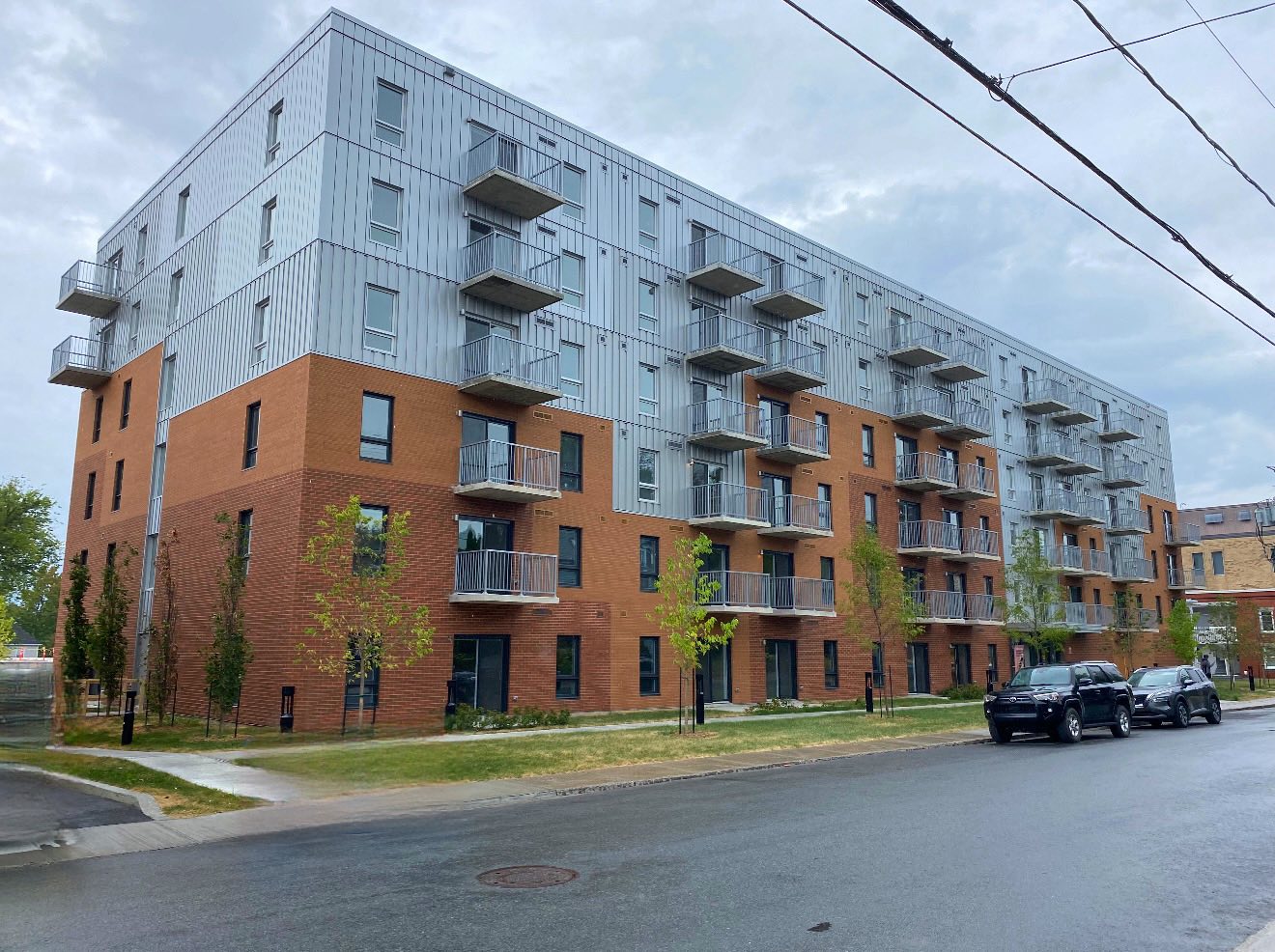Résidence Mission Unitaînés is a modern five-story apartment building with a brick lower facade and metal upper facade, featuring multiple balconies. Several trees line the sidewalk, and two parked cars are visible on the wet street in front.