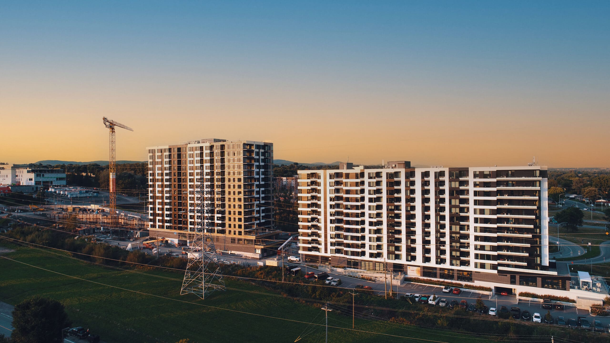 Vue aérienne de deux immeubles d'appartements modernes en construction, avec une grue sur le côté gauche, des lignes électriques au premier plan et une zone herbeuse. Le ciel du coucher de soleil projette une lueur chaleureuse sur les bâtiments et les environs.