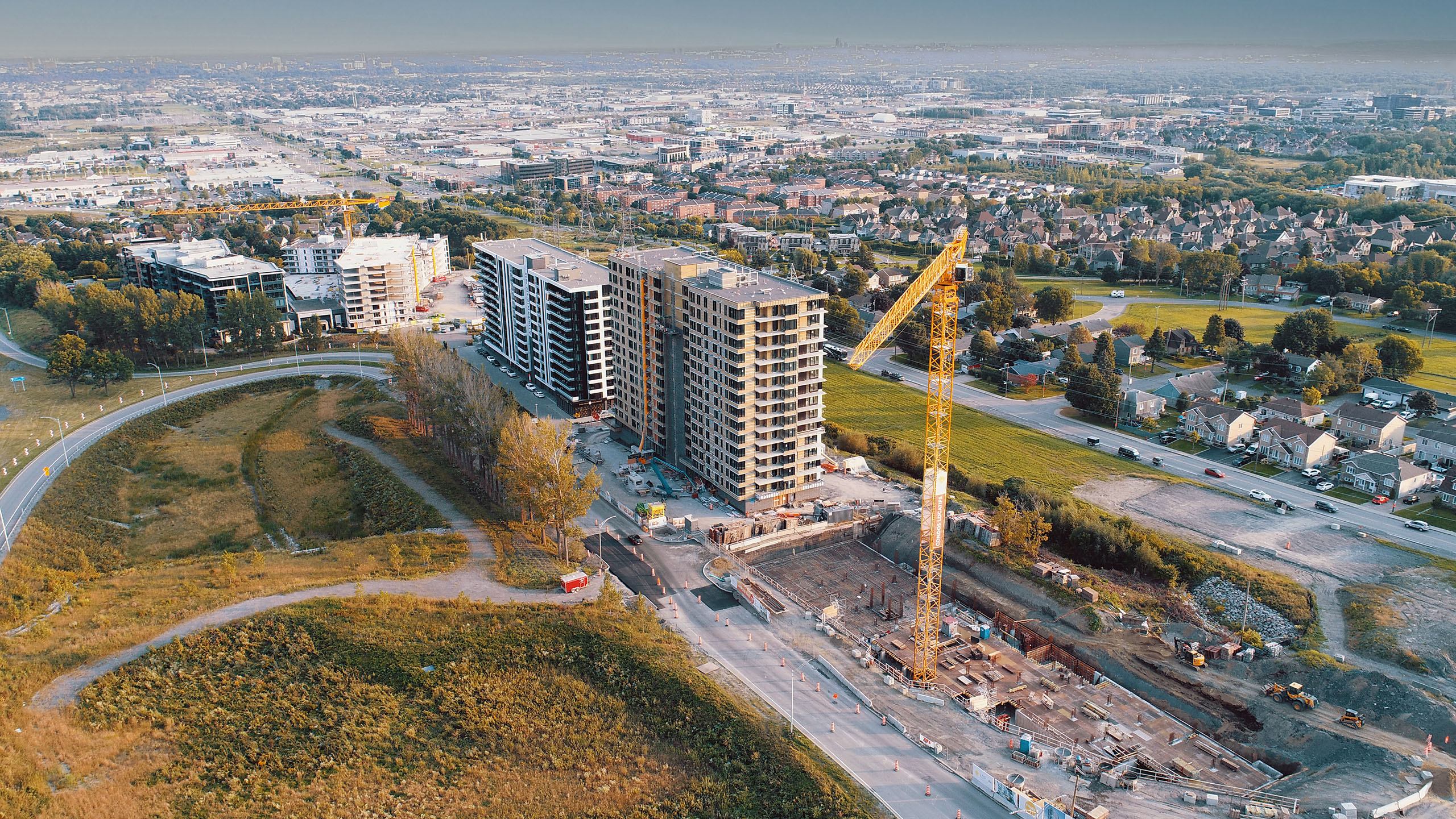 Vue aérienne d'un chantier de construction avec plusieurs immeubles de grande hauteur en cours de construction. Une grande grue est visible et divers matériaux et équipements de construction sont posés au sol. Autour du chantier se trouvent des quartiers résidentiels, des espaces verts et des infrastructures urbaines.