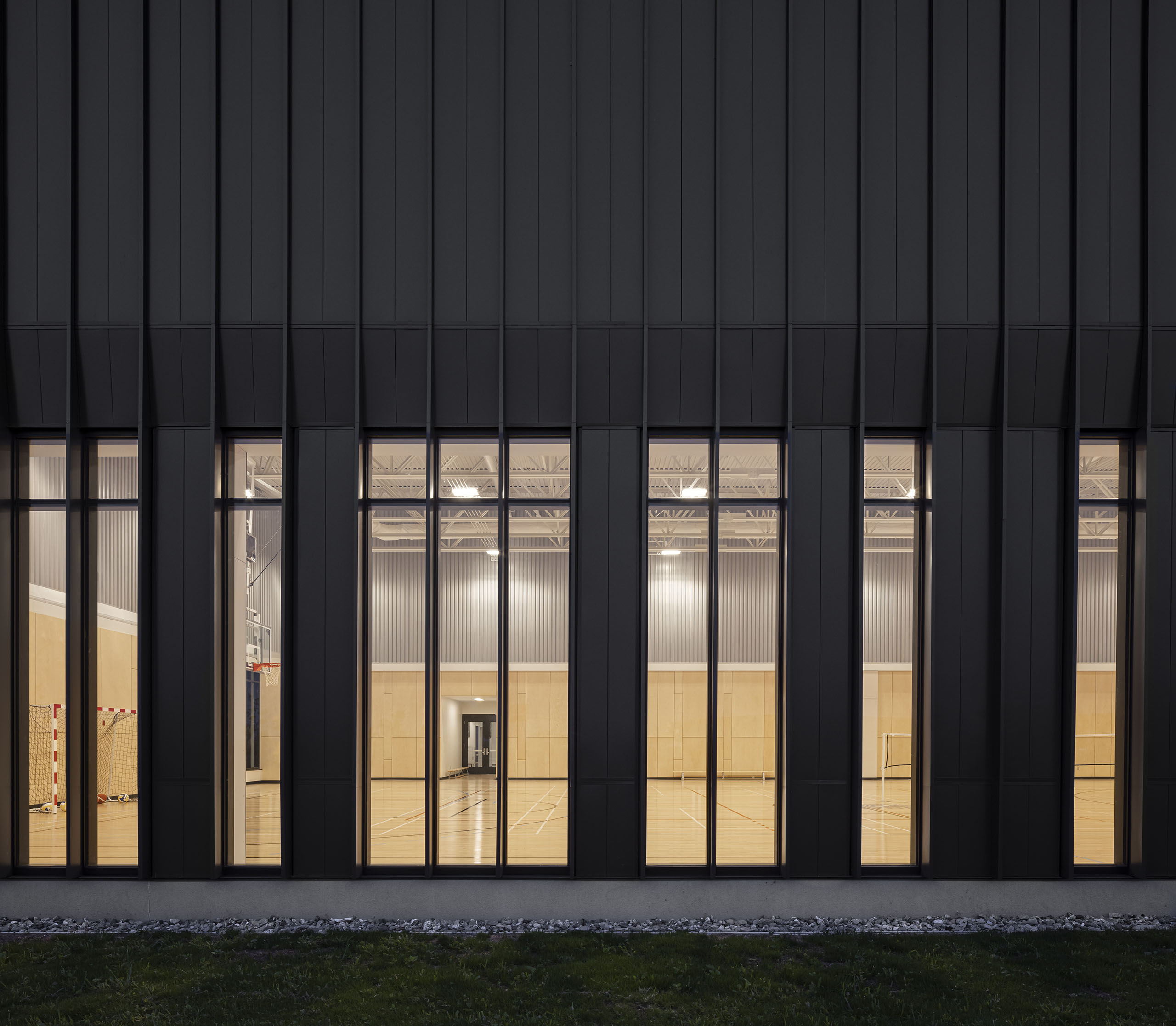 Vue extérieure d'un complexe sportif moderne avec de grandes fenêtres verticales, révélant un gymnase intérieur très éclairé avec des murs en bois et des paniers de basket. Le bâtiment présente une façade sombre et une bordure de gravier à la base avec de l'herbe au premier plan.