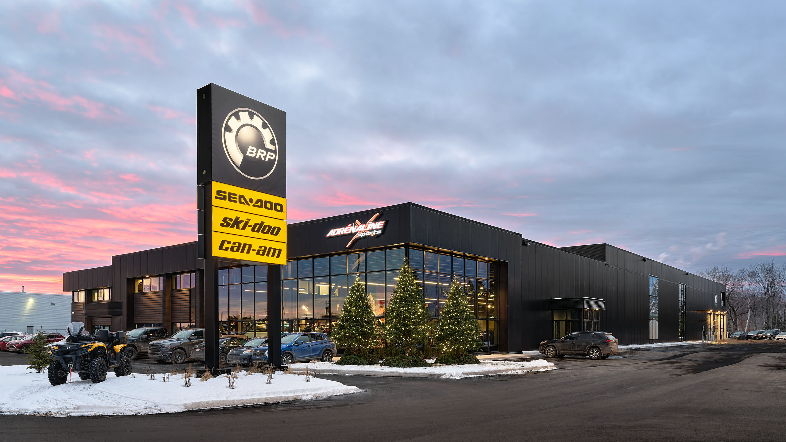 Un bâtiment moderne et élégant avec de grandes fenêtres en verre se présente au crépuscule sur un magnifique ciel rose et bleu. Le nouveau bâtiment est orné d'arbres de Noël et de lumières. Un panneau bien visible affiche les logos des marques BRP, Sea-Doo, Ski-Doo et Can-Am.