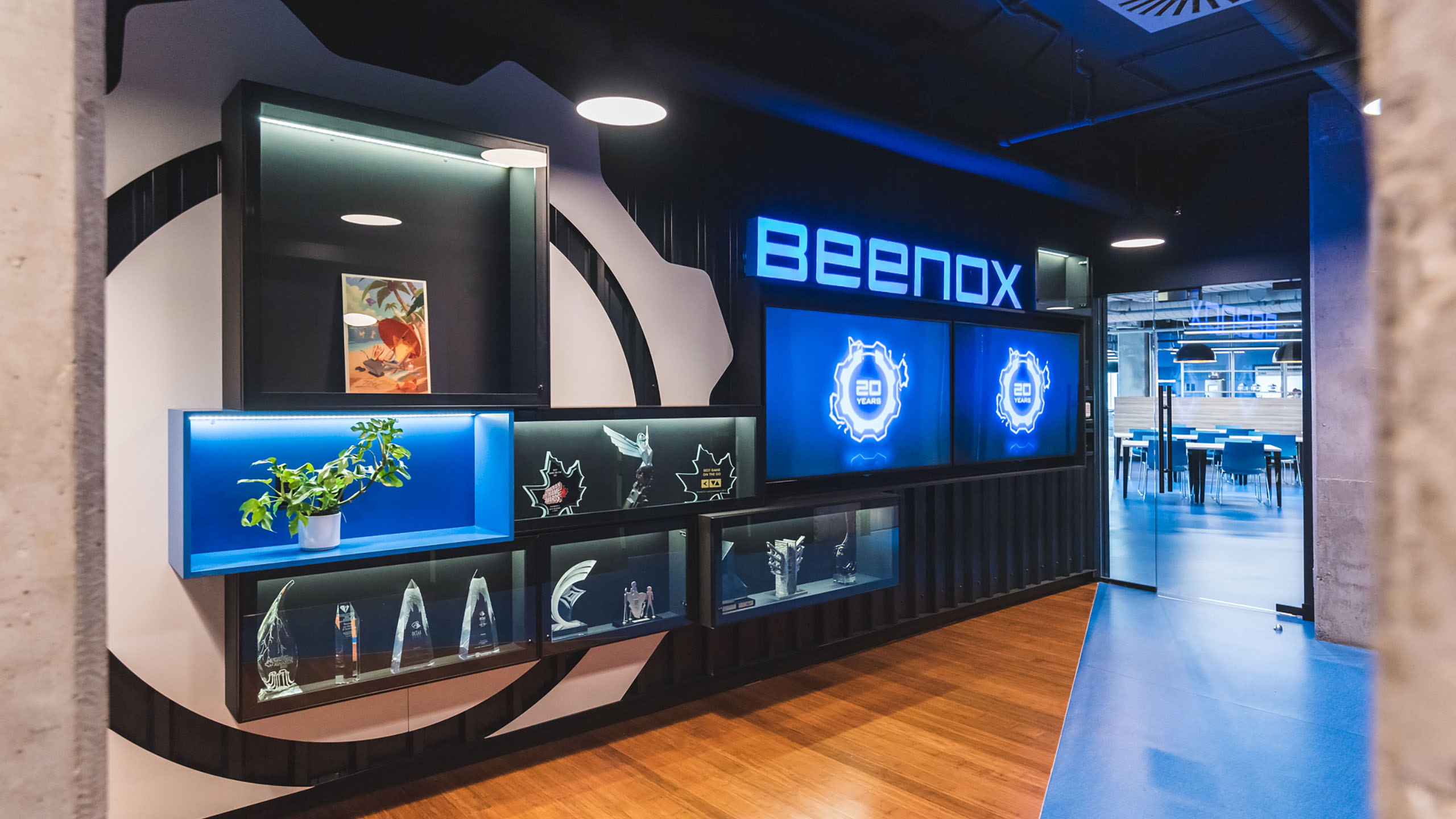 Un hall d'entrée de bureaux moderne au décor bleu et noir reflète la récente rénovation des espaces bureaux. Le mur affiche le logo lumineux « Beenox » et plusieurs étagères de vitrine éclairées contenant des trophées, des récompenses et une plante en pot. Une œuvre d'art encadrée est également visible sur le mur. Un aperçu d'une autre pièce est visible à travers une porte vitrée.