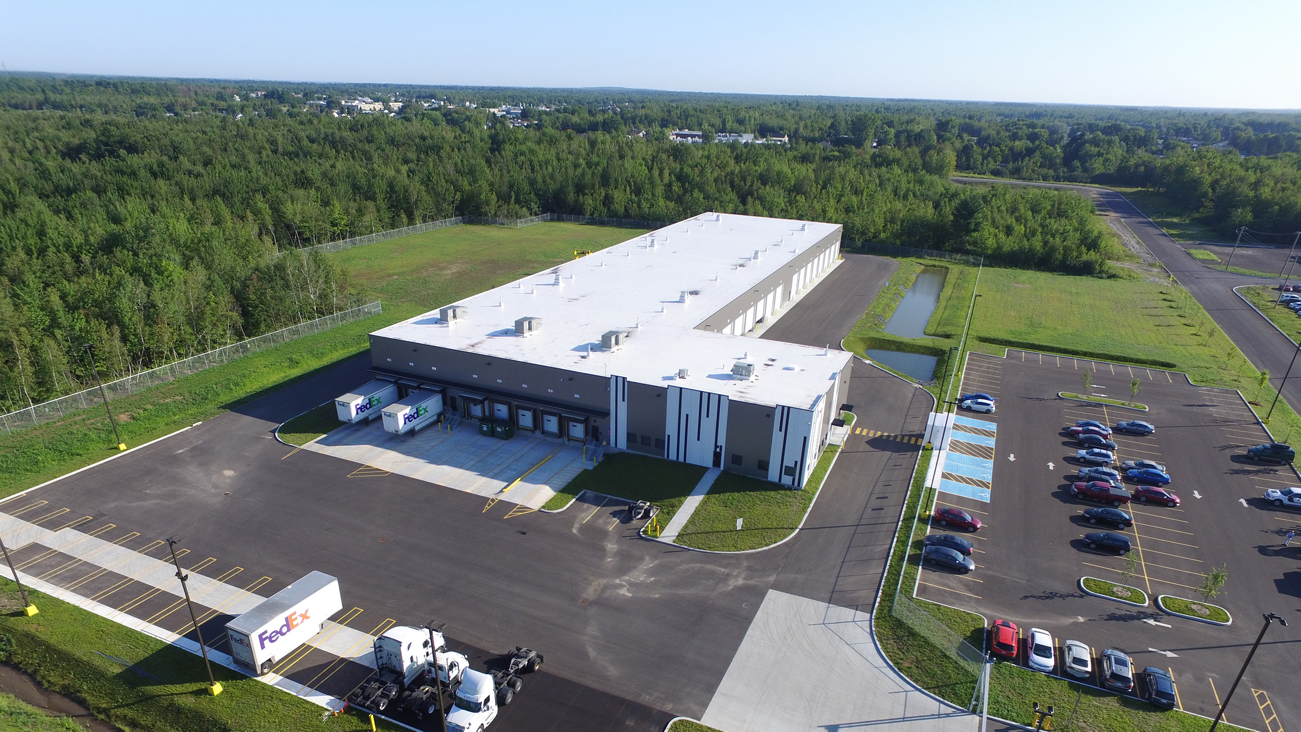 Vue aérienne d'un grand entrepôt FedEx de plain-pied au toit blanc, entouré d'un vaste parking. La construction d'un centre de distribution se déroule sur un fond d'arbres verts luxuriants et d'espaces verts, avec plusieurs voitures garées et camions de livraison visibles.