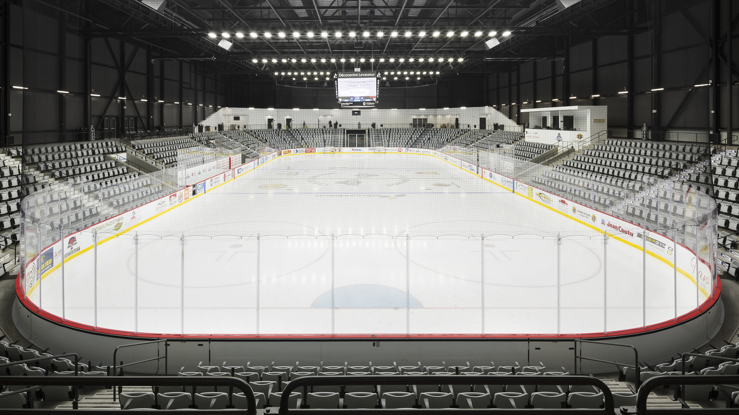 Vue grand angle d'une patinoire de hockey sur glace intérieure vide avec des marquages rouges et jaunes, entourée de sièges gris vides dans les gradins. Des lumières au plafond illuminent la patinoire comme un amphithéâtre méticuleux, et un grand tableau d'affichage est suspendu au-dessus d'une extrémité, n'affichant aucun contenu actif pendant que la construction se poursuit.