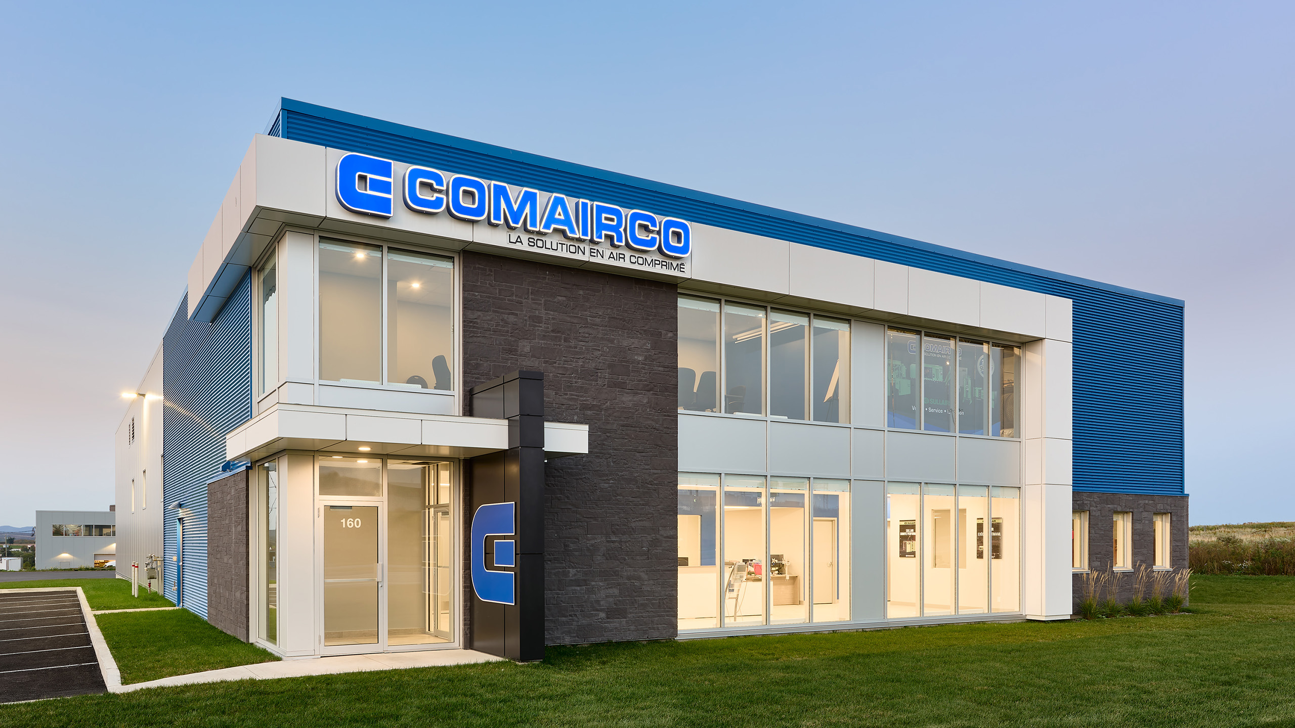 Un bâtiment commercial moderne de deux étages avec de grandes fenêtres en verre et un extérieur aux tons bleu, blanc et gris. Un panneau bien visible indique « Comairco » avec le texte français « La Solution En Air Comprimé » sur la façade. Conçu avec une attention particulière portée à la conception et à la construction, il dispose d'une pelouse verte à l'avant et d'un parking à proximité.