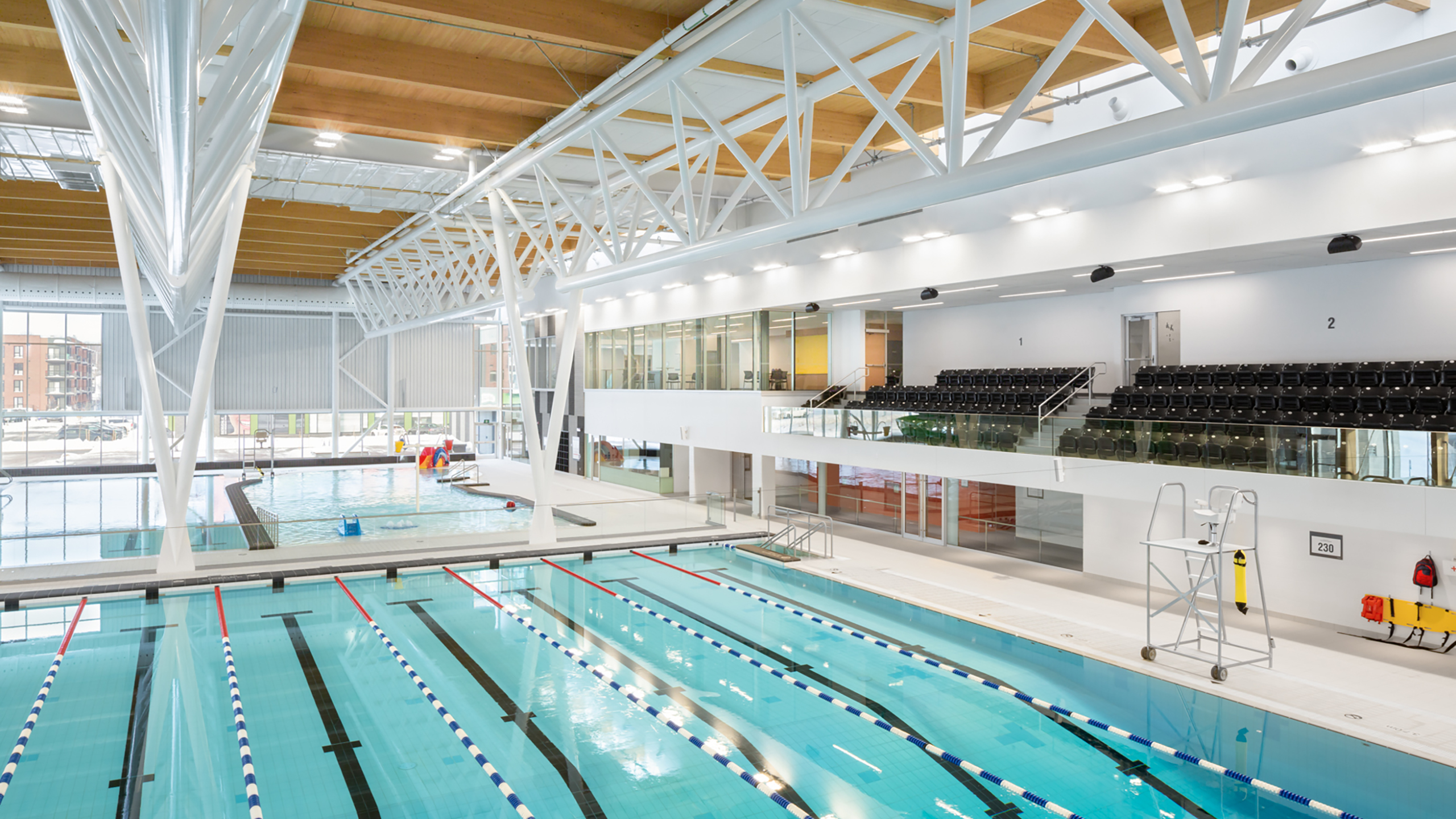 Une piscine intérieure moderne avec plusieurs couloirs de nage, des blocs de départ et un bassin bleu clair. La construction d'un bâtiment multifonctionnel comprend un haut plafond avec des poutres blanches apparentes, des sièges pour les spectateurs et de grandes fenêtres permettant à la lumière naturelle de remplir l'espace.