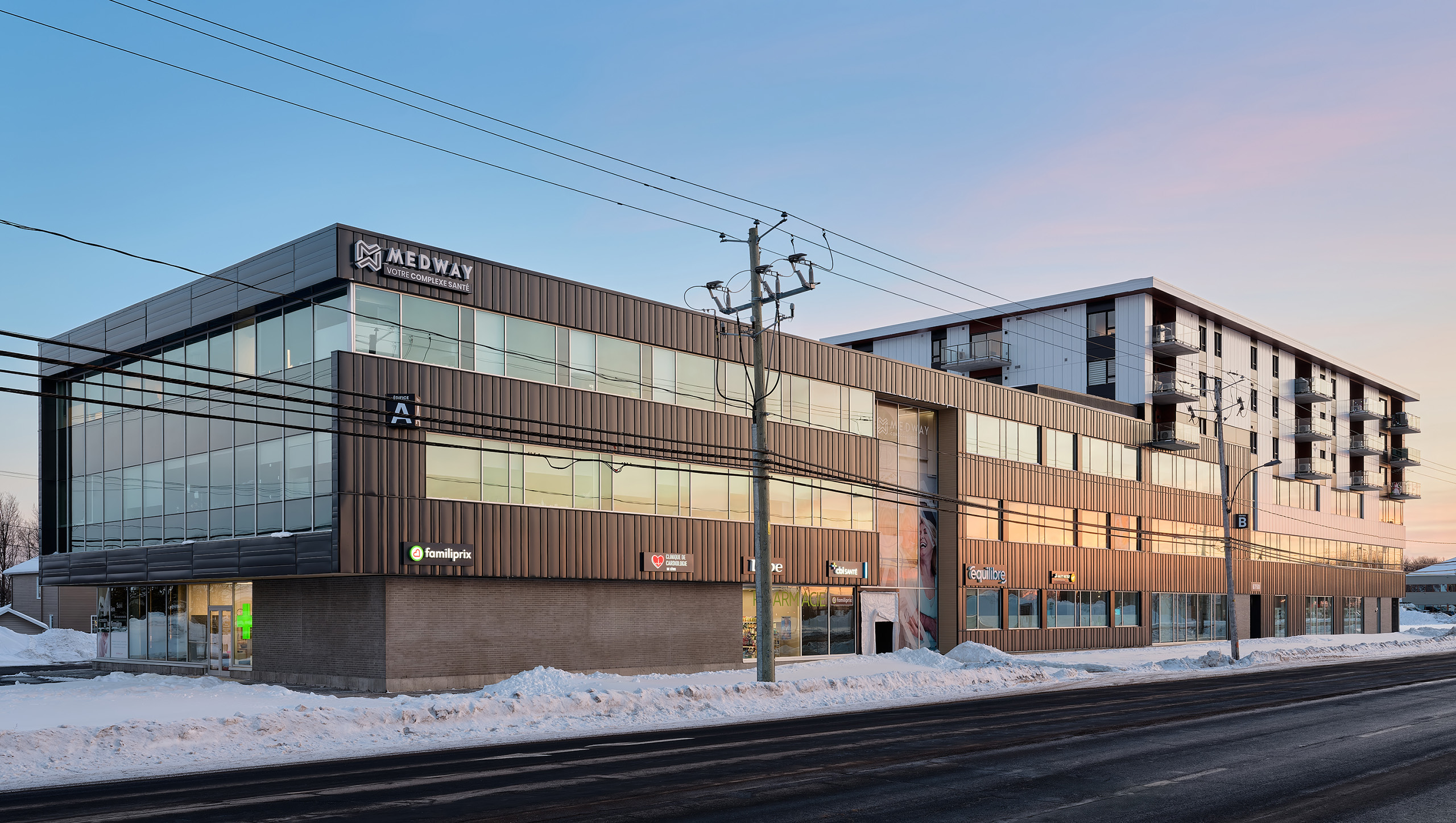 Un bâtiment moderne de trois étages avec de grandes fenêtres en verre et des panneaux métalliques sombres à l'extérieur, présentant des enseignes pour diverses entreprises, dont Medway. La neige recouvre le sol tandis que des lignes électriques traversent le ciel. La scène, qui se déroule au coucher du soleil avec un ciel clair, laisse entrevoir un agrandissement majeur en cours.