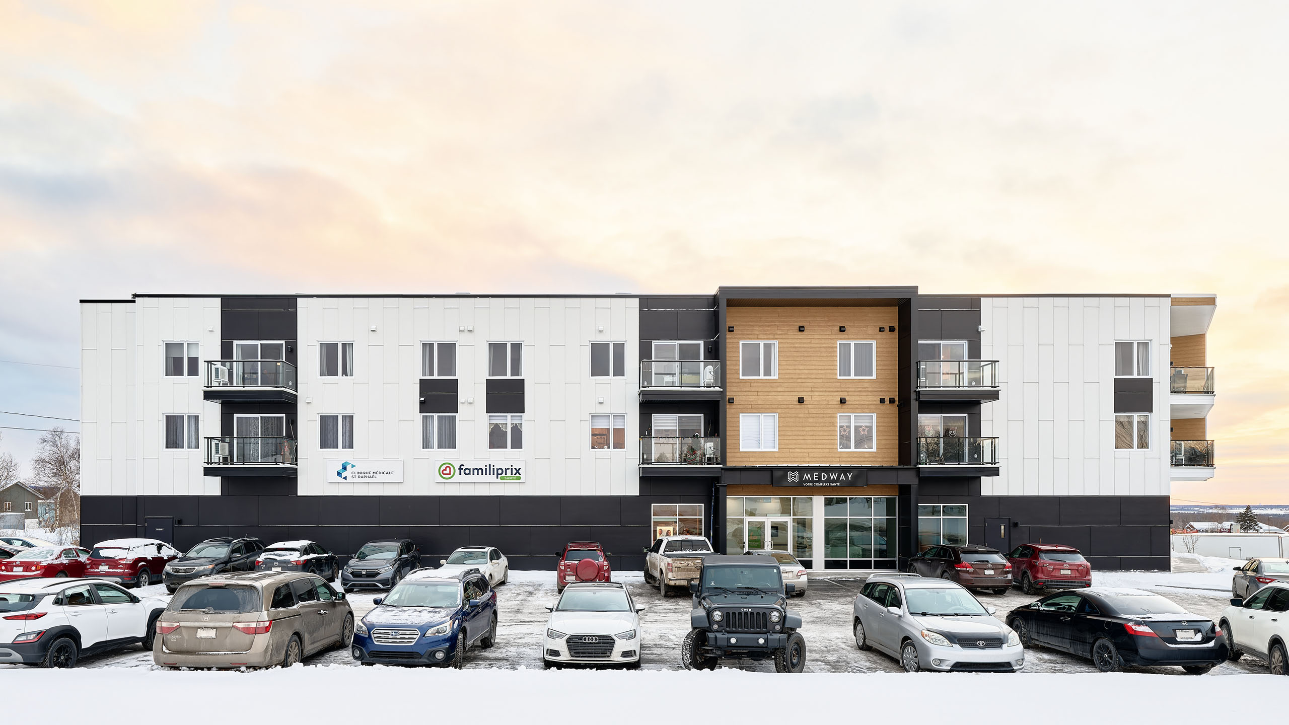 Un immeuble d'appartements moderne de trois étages avec une façade en lambris de bois, de noir et de blanc se dresse au milieu des travaux de construction d'un nouveau bâtiment. Plusieurs voitures sont garées sur le terrain enneigé en face, tandis que des panneaux pour « Familiprix » et d'autres commerces sont visibles au rez-de-chaussée.