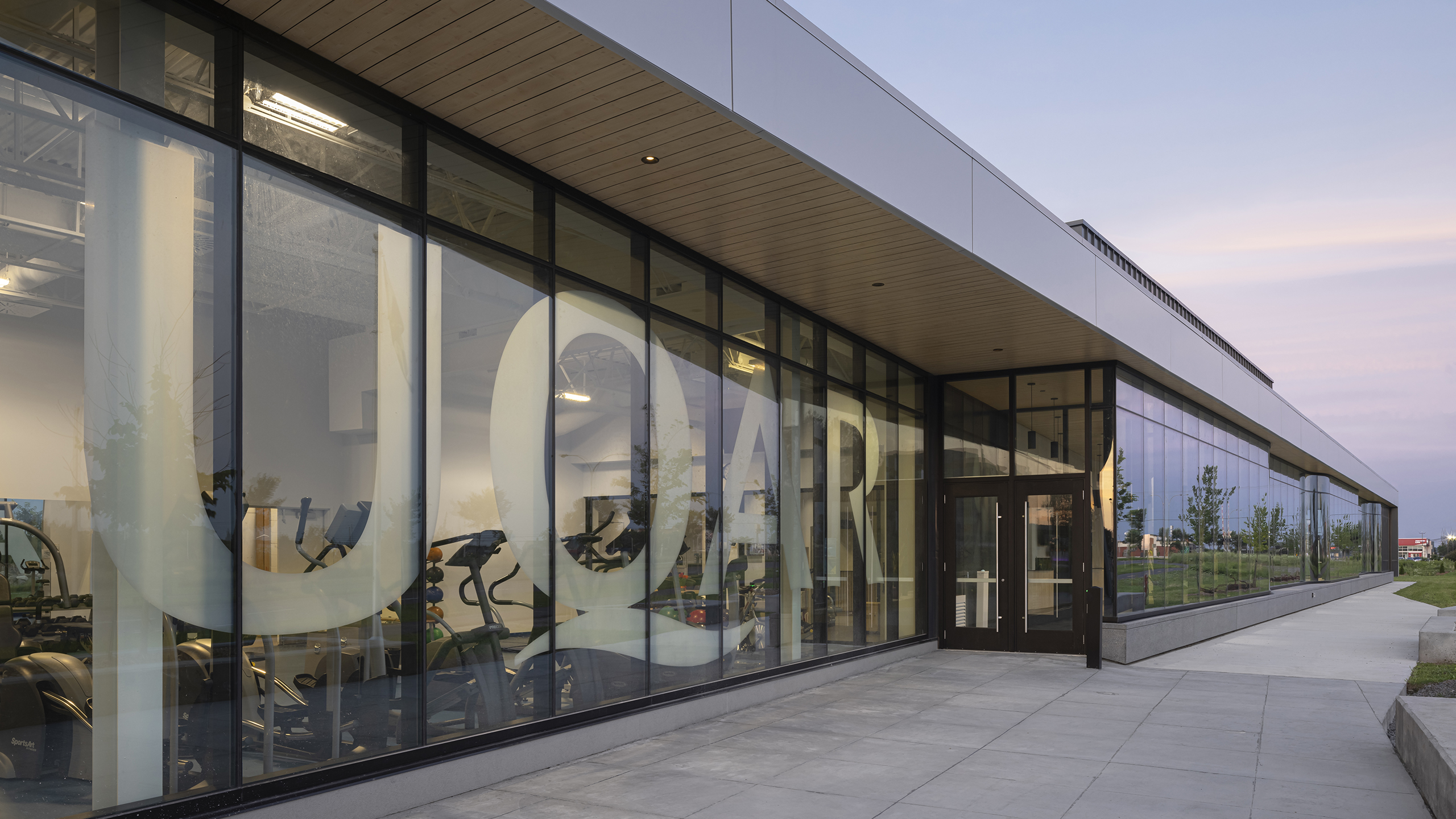 Vue extérieure d'un bâtiment moderne avec de grandes baies vitrées exposant des équipements d'entraînement à l'intérieur, tels que des vélos stationnaires et des vélos elliptiques. Le mot « UQAM » est affiché en évidence sur les fenêtres. Le ciel apparaît serein avec des couleurs claires, indiquant le début de soirée ou le matin, un bel exemple de construction d'un complexe sportif.