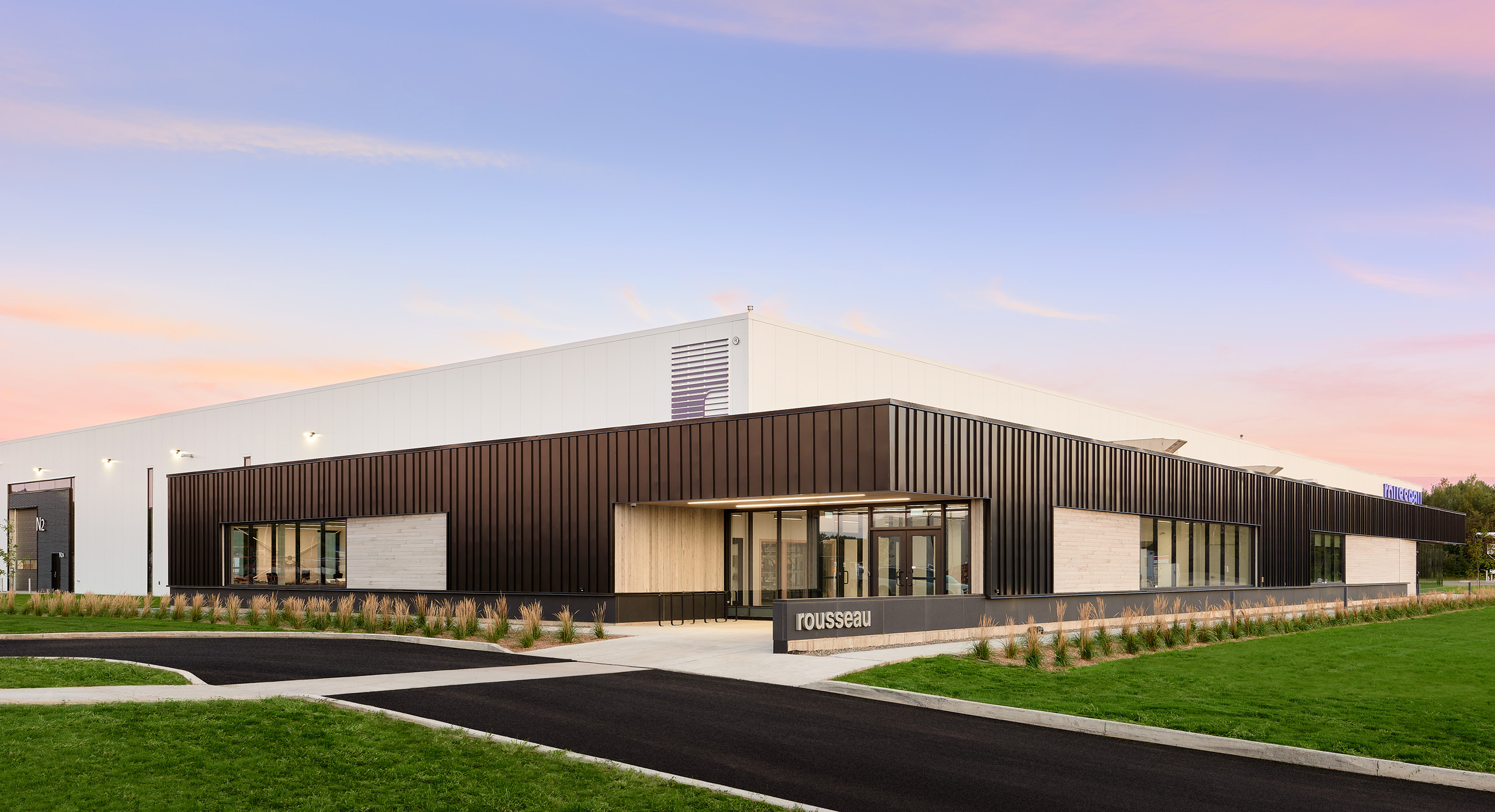 Un bâtiment moderne de plain-pied avec un toit plat et une combinaison de murs extérieurs blancs et brun foncé. La façade avant est dotée de grandes fenêtres et d'une entrée principale, entourée de pelouses paysagères et de sentiers. "Rousseau" est écrit près de l'entrée, marquant la construction d'une nouvelle usine.