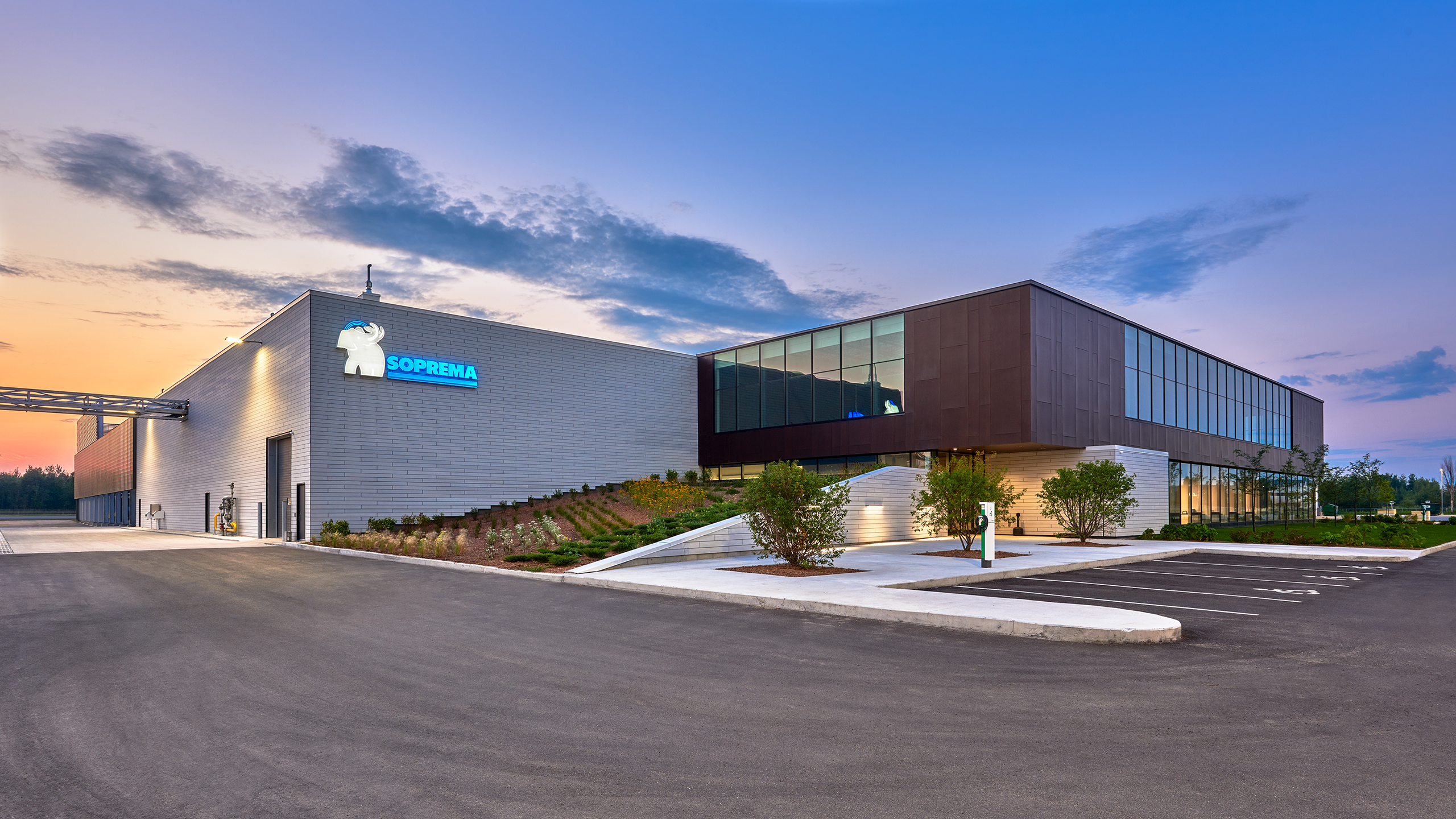 Un grand entrepôt moderne est visible au crépuscule avec un extérieur en brique et en verre. La construction nouvelle usine porte le logo « Soprema » avec un bélier dans le coin supérieur. Un petit parking et des espaces verts paysagers se trouvent devant la structure. Le ciel est clair avec un dégradé du bleu clair au bleu plus foncé.