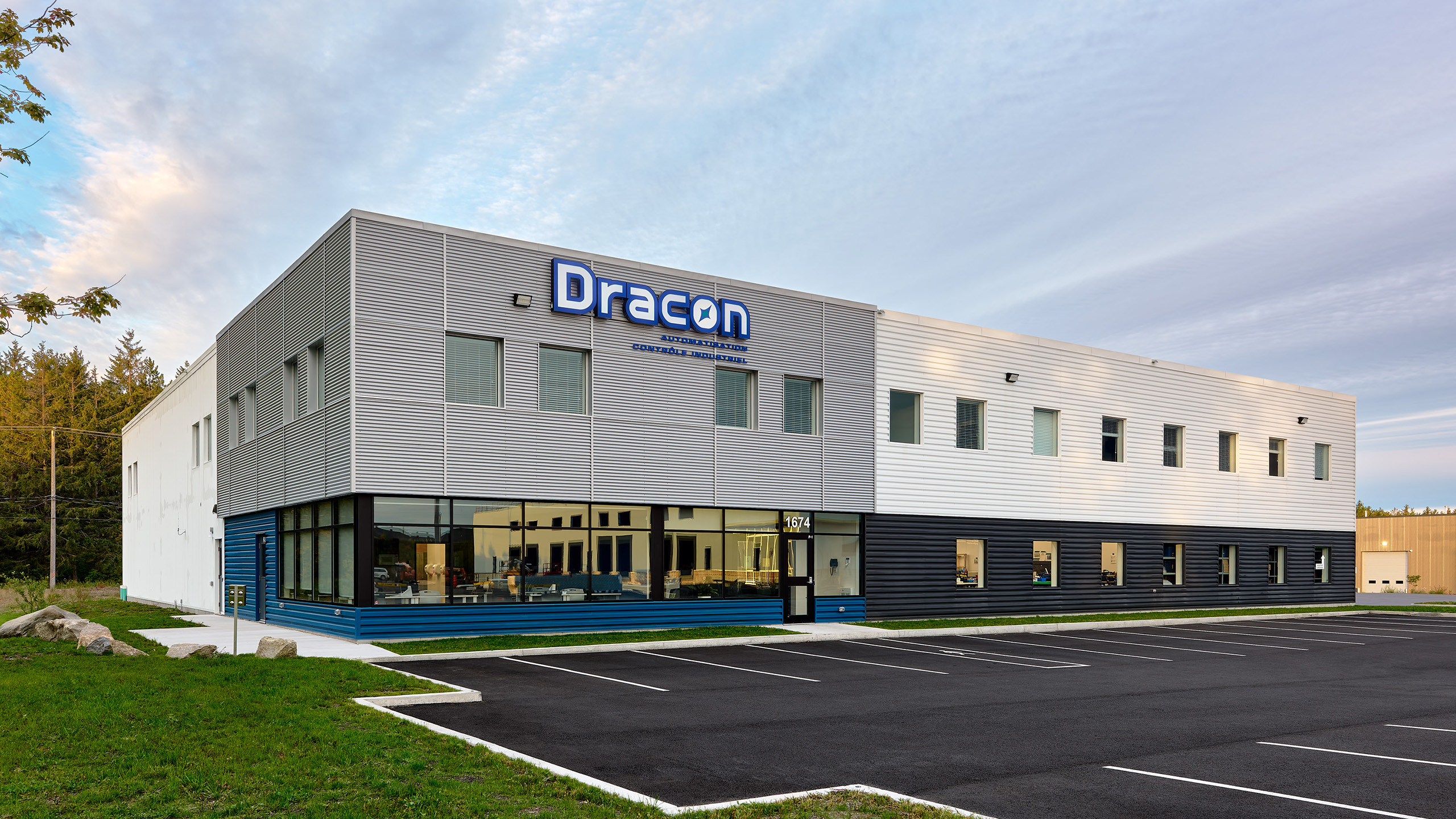 Un bâtiment commercial moderne de deux étages étiqueté « Dracon » avec « Manufacturing Excellence » sous le nom. La façade combine des panneaux gris, blancs et bleus avec de nombreuses fenêtres, mettant en valeur la construction d'un nouveau bâtiment. Le bâtiment fait face à un parking vide sous un ciel partiellement nuageux.