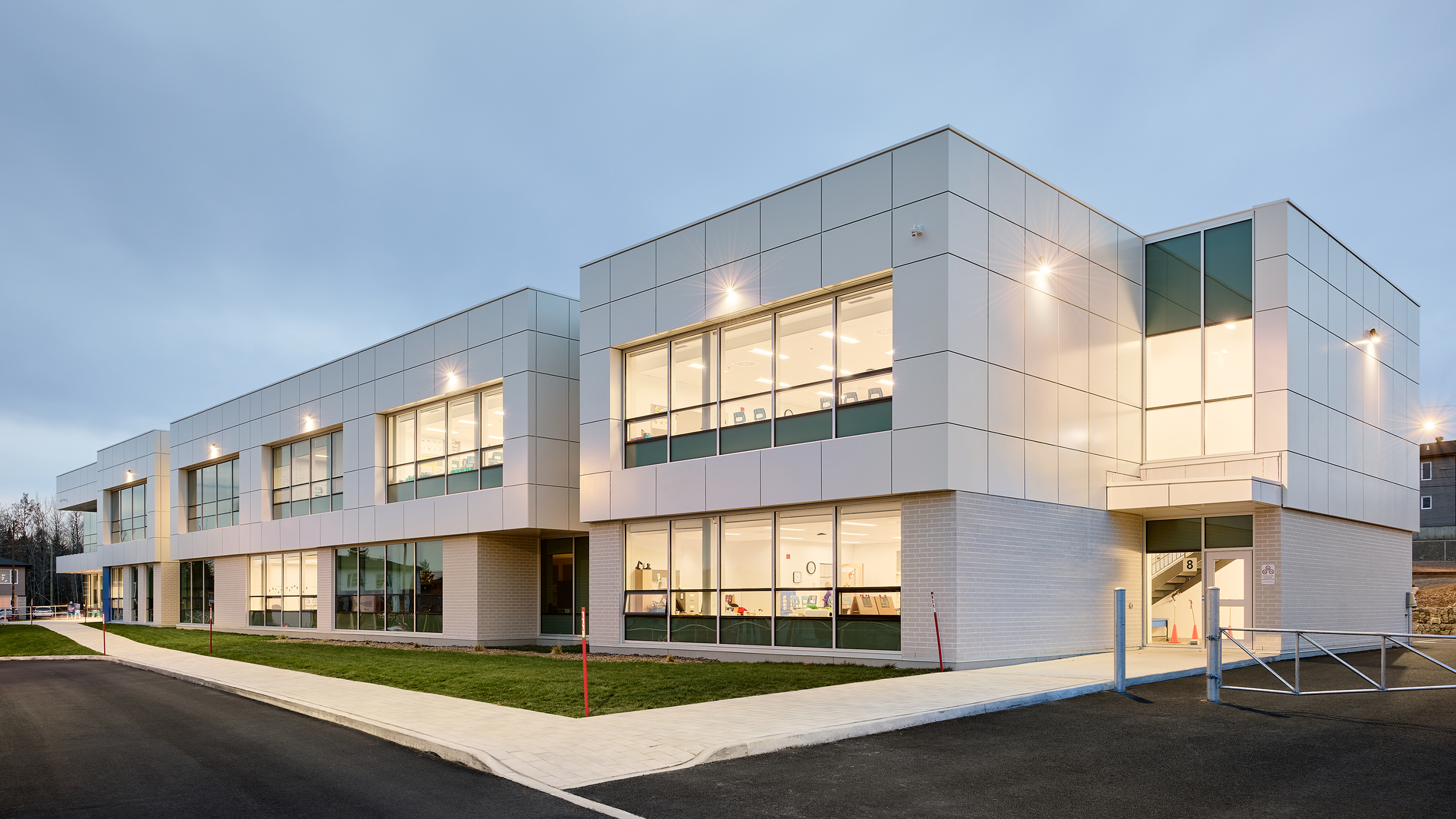 Un bâtiment commercial moderne de deux étages au design épuré avec de grandes fenêtres et des panneaux blancs. L'extérieur, semblable à celui de la construction d'une nouvelle école, comprend une allée bien éclairée et une petite zone gazonnée. Le ciel est couvert, ce qui indique qu'il est tôt le matin ou en fin d'après-midi.