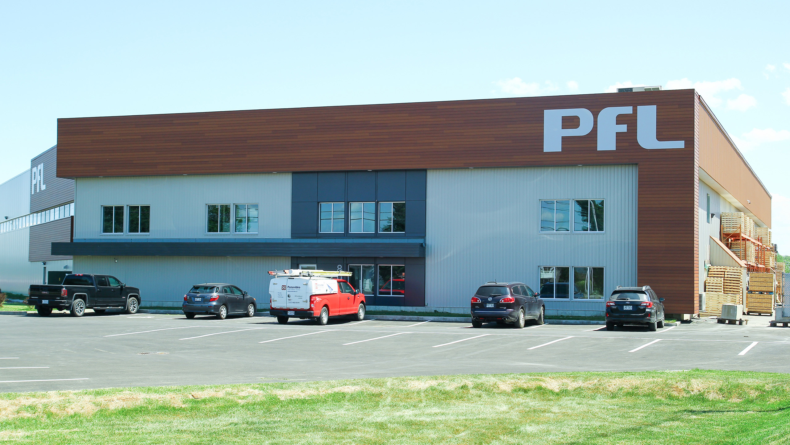 Un bâtiment commercial moderne de deux étages avec un mélange de bois et de métal, portant les grandes lettres blanches « PFL » dans le coin supérieur droit, symbolise l’agrandissement récent d’une usine existante. Plusieurs voitures et une camionnette sont stationnées devant sur un terrain pavé avec une pelouse bien entretenue au premier plan.
