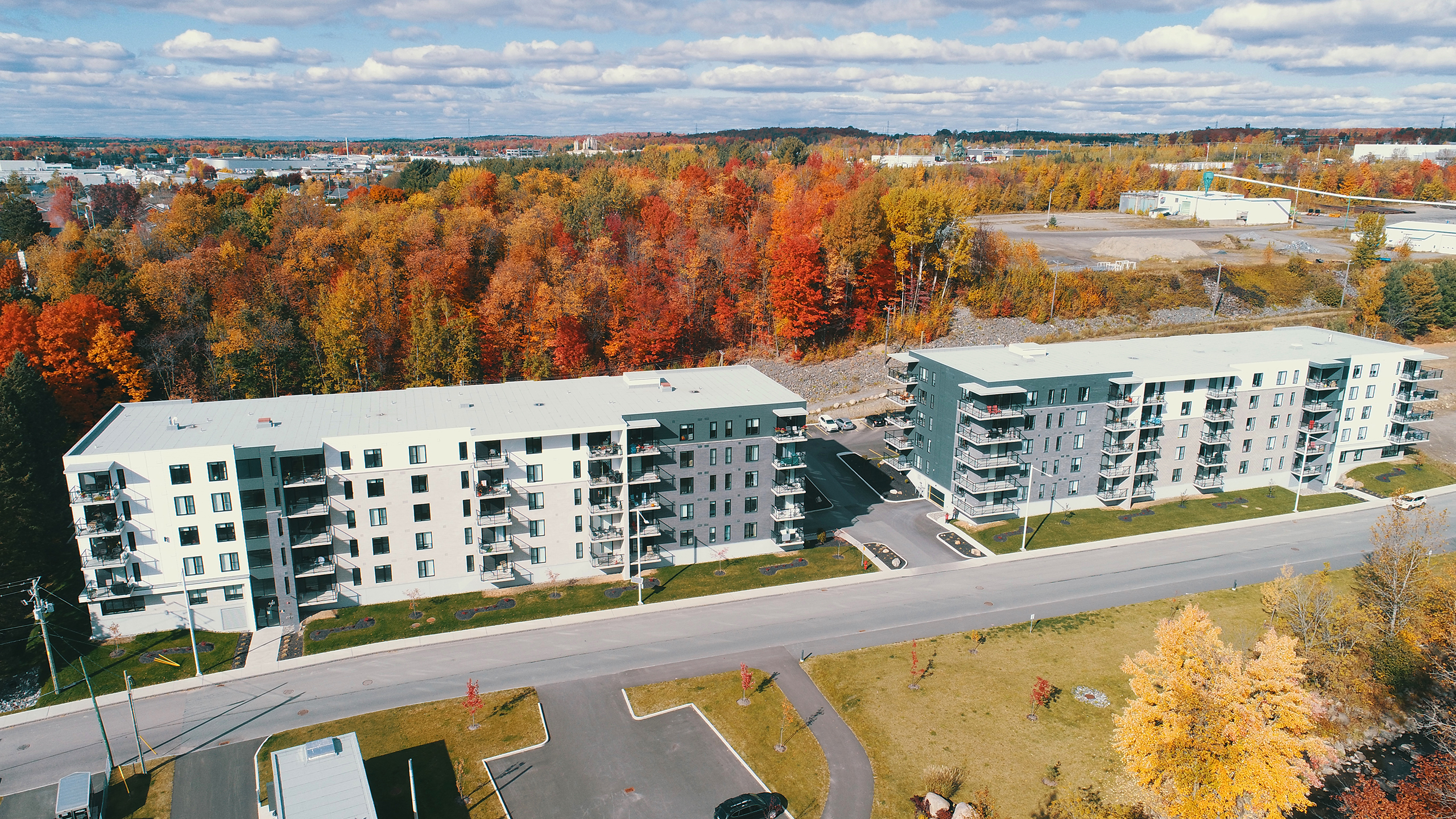 Vue aérienne de deux immeubles d'appartements modernes à plusieurs étages présentant une impressionnante construction multirésidentielle, entourée d'un feuillage d'automne vibrant. Une route passe entre les bâtiments, avec des parcelles d'arbres verts et colorés autour. Le ciel est partiellement nuageux avec un paysage lointain à l'horizon.