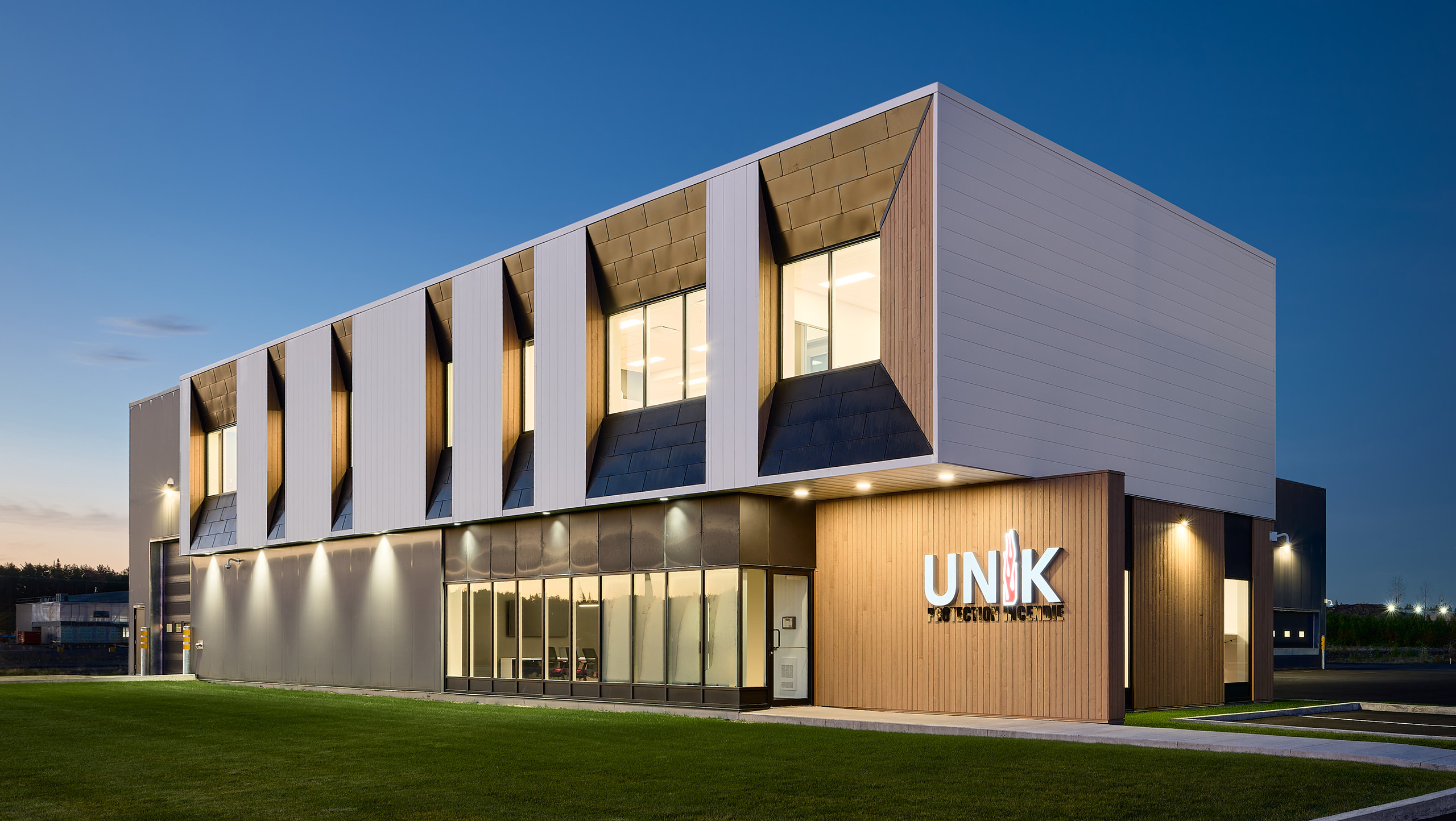 Un bâtiment moderne de deux étages au design architectural épuré, doté de grandes fenêtres et d'une combinaison de panneaux extérieurs blancs et gris. L'entrée arbore le mot « UNIK » bien en évidence. Cette construction du nouveau siège social est magnifiquement mise en valeur dans une scène de crépuscule avec des lumières illuminant la structure.