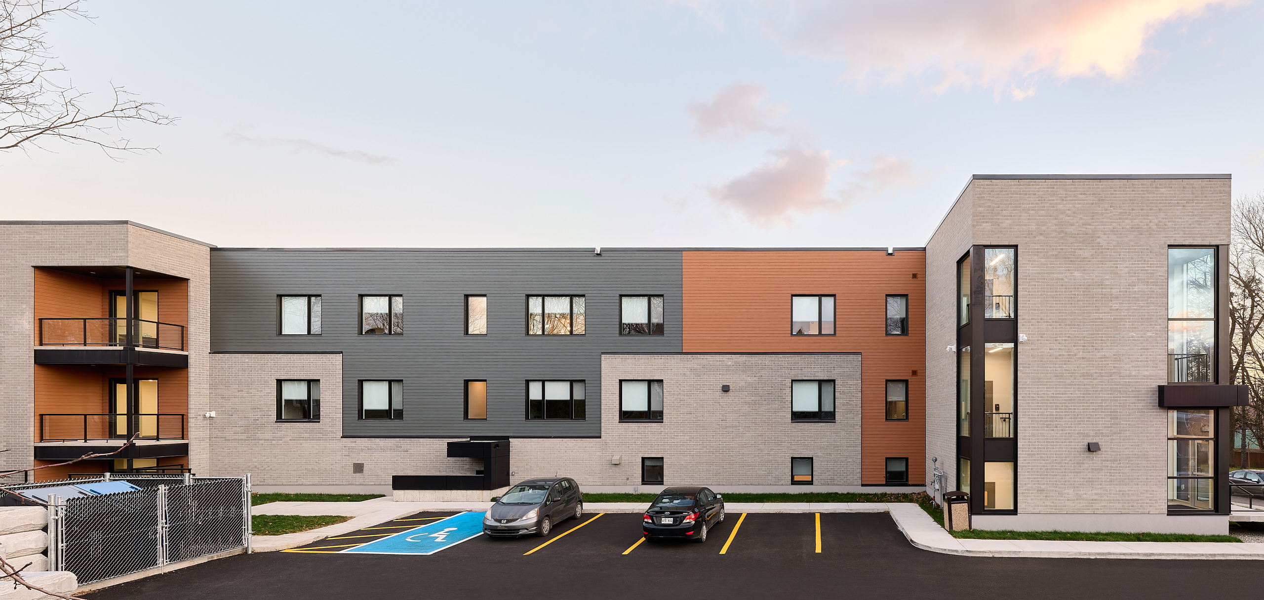 Un immeuble d'appartements moderne de trois étages avec un mélange de panneaux extérieurs gris, orange et beige est un exemple de construction récente d'un nouveau bâtiment. La structure dispose de grandes fenêtres et de balcons. Devant, il y a un parking avec deux voitures et une place de parking accessible marquée. Le ciel est partiellement nuageux.