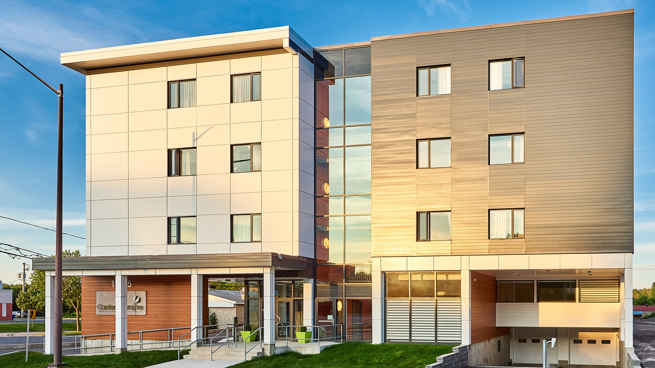 Une résidence moderne de quatre étages avec un mélange de panneaux extérieurs blancs et bruns, de grandes fenêtres et une section centrale en verre. L'entrée comprend une passerelle couverte avec des sièges, un aménagement paysager vert et un panneau indiquant « 5 ». Une allée et un parking sont visibles en contrebas.
