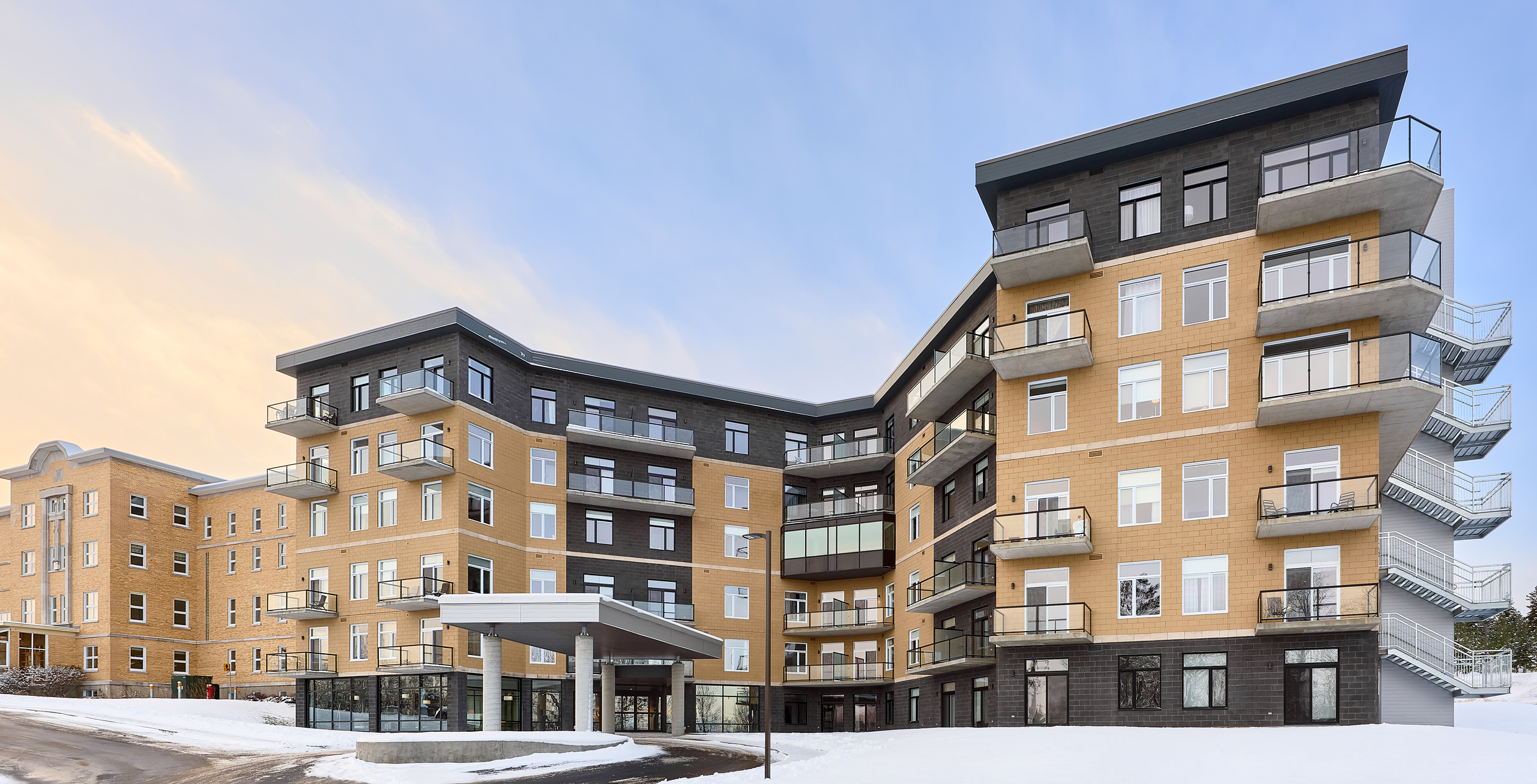 Un immeuble d'appartements moderne à plusieurs étages avec une combinaison d'extérieurs beiges et noirs. La structure dispose de nombreux balcons, de grandes fenêtres et d'une entrée couverte. La neige recouvre le sol et le ciel présente un doux dégradé de tons chauds et froids, parfait pour une construction de résidence pour personnes âgées à proximité.