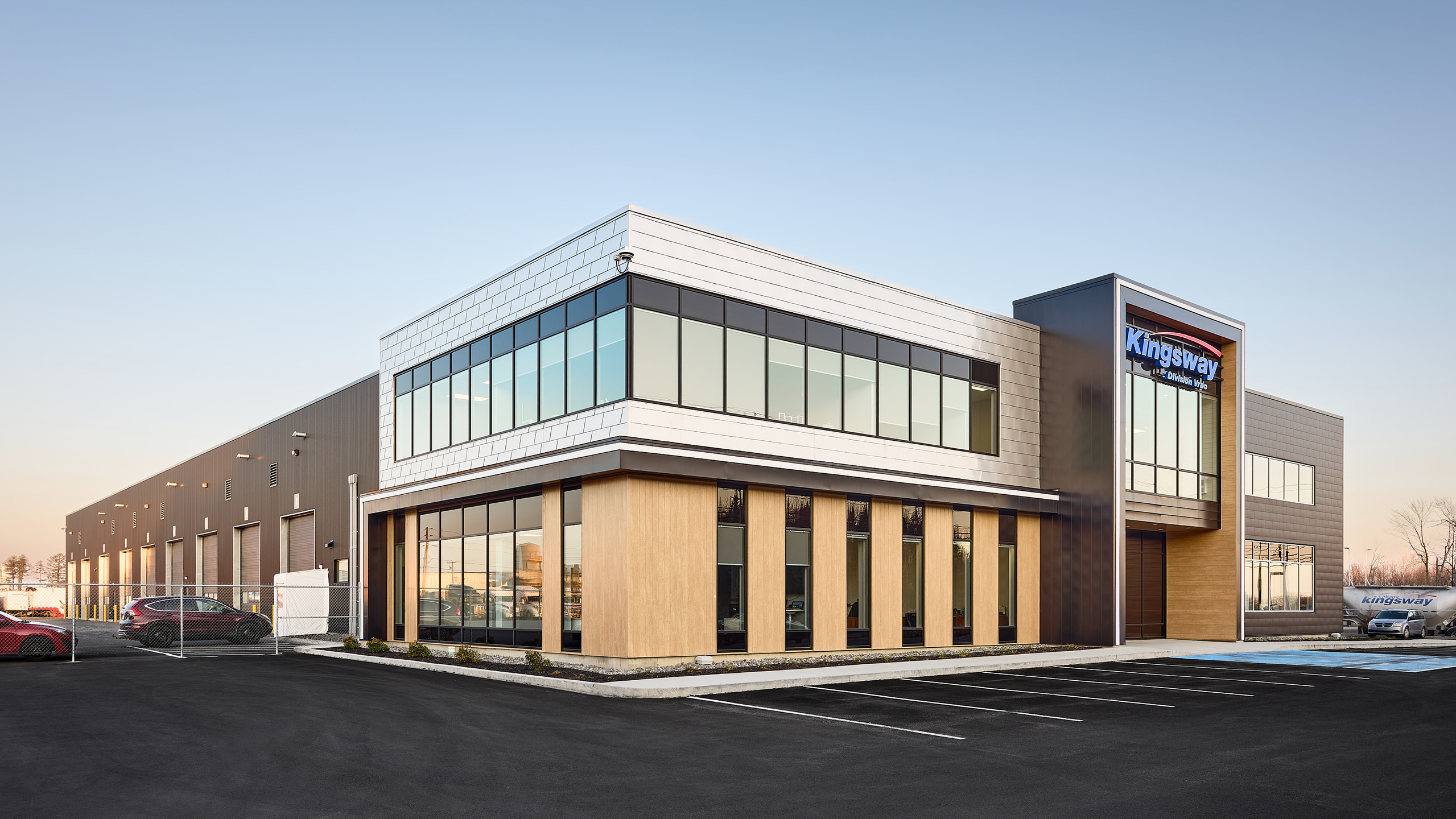 Un bâtiment commercial moderne de deux étages avec de grandes fenêtres et un extérieur aux tons gris et crème. Un logo indiquant « Kingsway » est affiché en évidence. Les environs comprennent un parking pavé avec plusieurs voitures garées et un ciel bleu clair au-dessus, faisant allusion à la récente construction d'un nouveau garage à proximité.