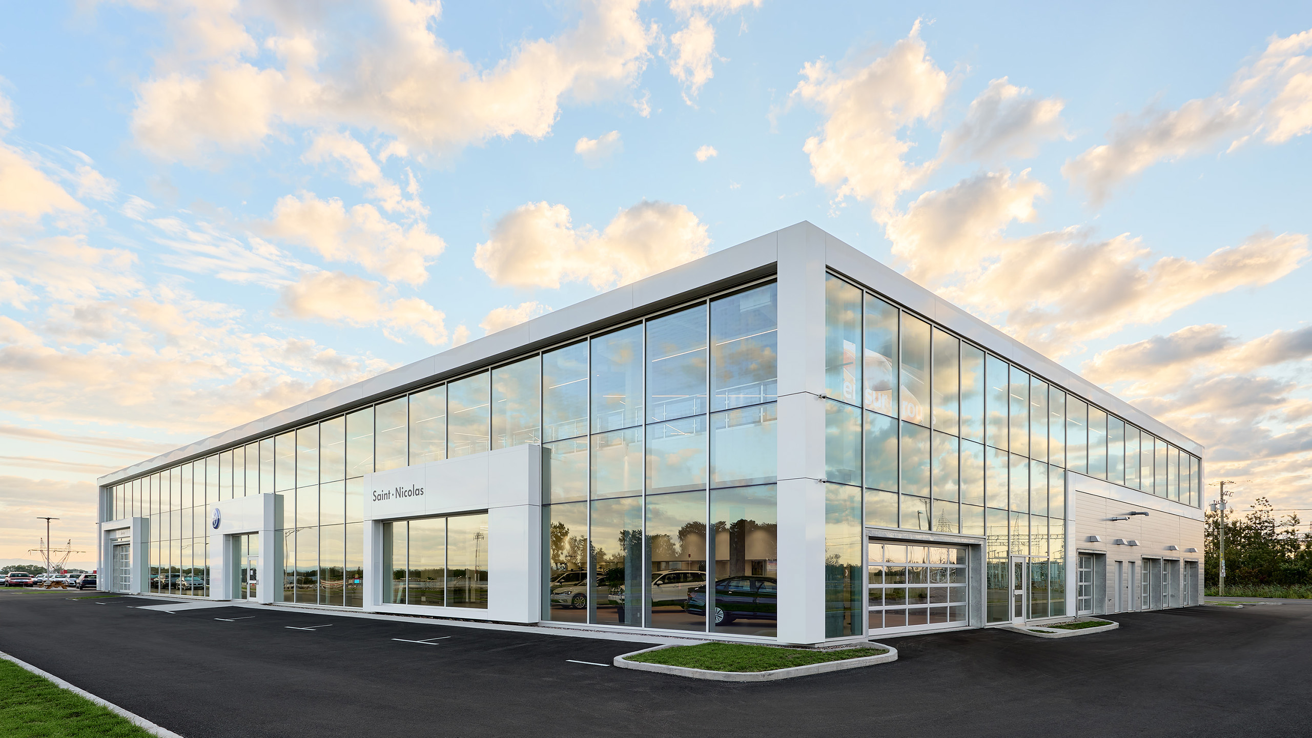 Un bâtiment moderne en verre et blanc avec de grandes fenêtres reflétant le ciel, avec des panneaux d'entrée « Ventes » et « Service », met en valeur la construction d'un nouveau concessionnaire. Le ciel est partiellement nuageux avec un mélange de nuages clairs et plus sombres, et les environs disposent d'une allée et d'un espace de stationnement bien entretenus.