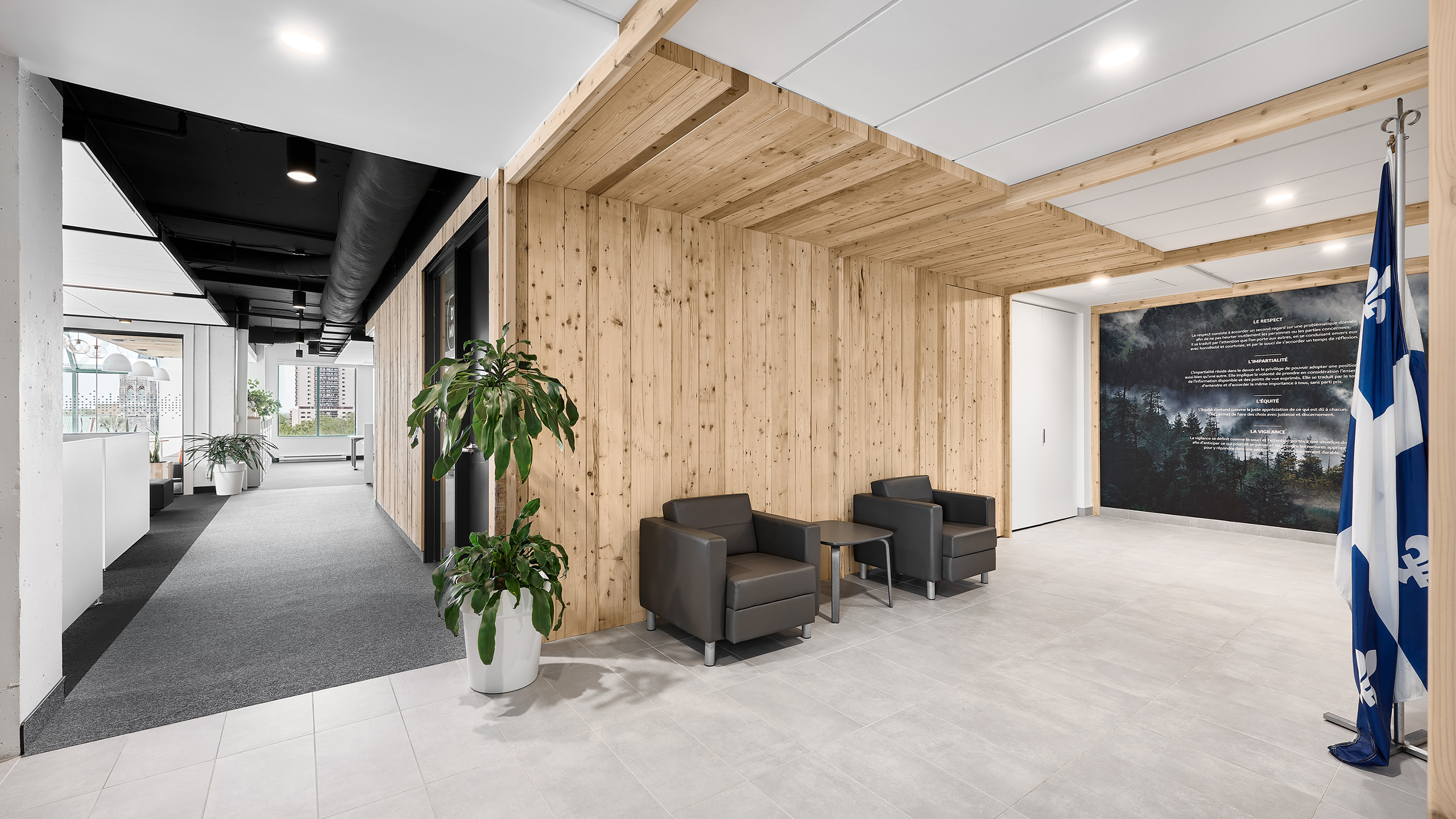Un espace de bureau moderne avec des murs en panneaux de bois, un sol en carrelage gris et une salle d'attente avec deux fauteuils gris foncé et une petite table. L'aménagement intérieur des bureaux comprend des plantes en pot, un drapeau du Québec et, en arrière-plan, un couloir avec un plafond noir et de grandes fenêtres.