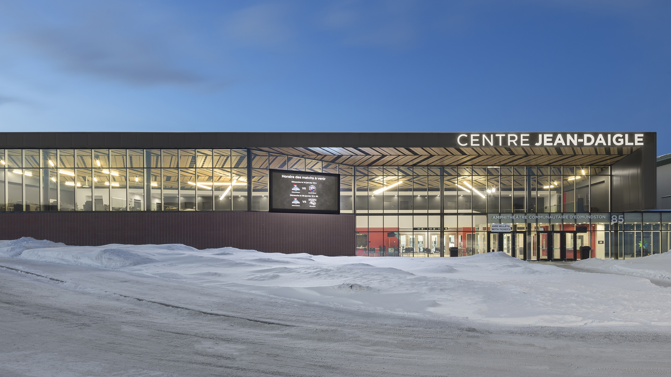 L'image montre l'extérieur du Centre Jean-Daigle, une construction moderne dotée de grandes fenêtres vitrées et d'un toit plat. Elle est entourée de neige, signe d'un climat froid. Le nom du bâtiment est affiché en évidence sur la façade, et un panneau d'affichage numérique complète cet amphithéâtre contemporain.
