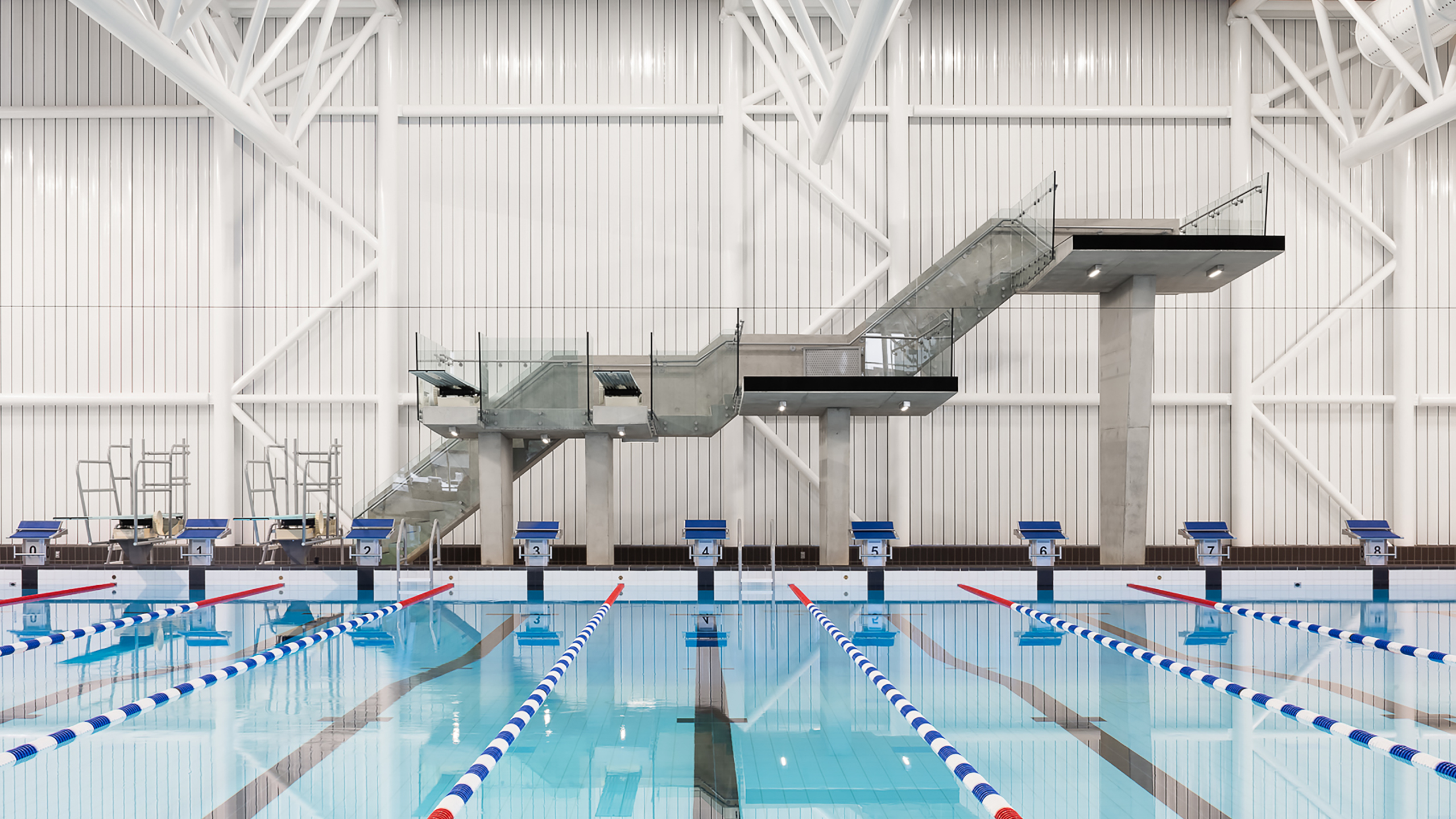 Piscine intérieure à couloirs multiples séparés par des séparations bleues et blanches. En arrière-plan, on distingue un ensemble de plongeoirs et de plateformes à différentes hauteurs, accessibles par un escalier. L'espace piscine, qui fait partie de la construction d'un bâtiment multifonctionnel, est entouré d'une structure blanche de style industriel.