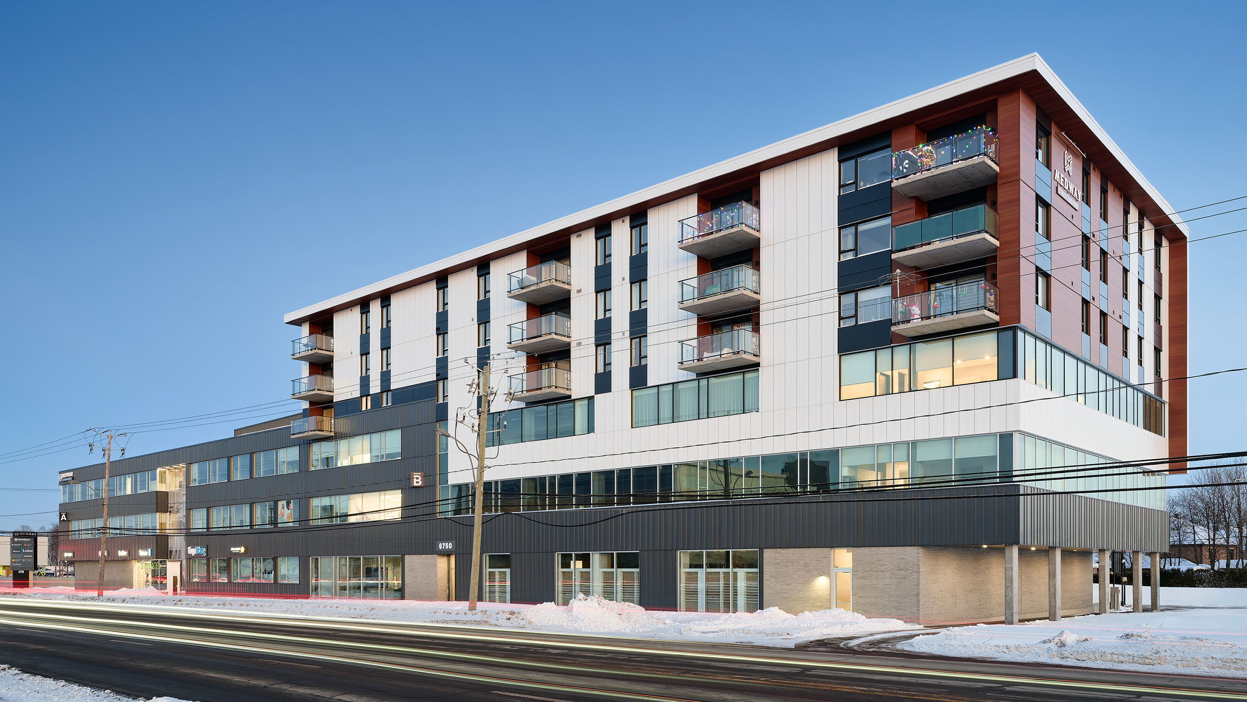 Un bâtiment moderne à plusieurs étages avec de grandes fenêtres et des balcons se dresse dans une rue bordée de neige. La structure, bénéficiant d'un agrandissement majeur, présente un mélange de panneaux extérieurs blancs, noirs et en bois. Des traînées lumineuses provenant des véhicules qui passent sont visibles à l'avant et des câbles électriques traversent la scène.