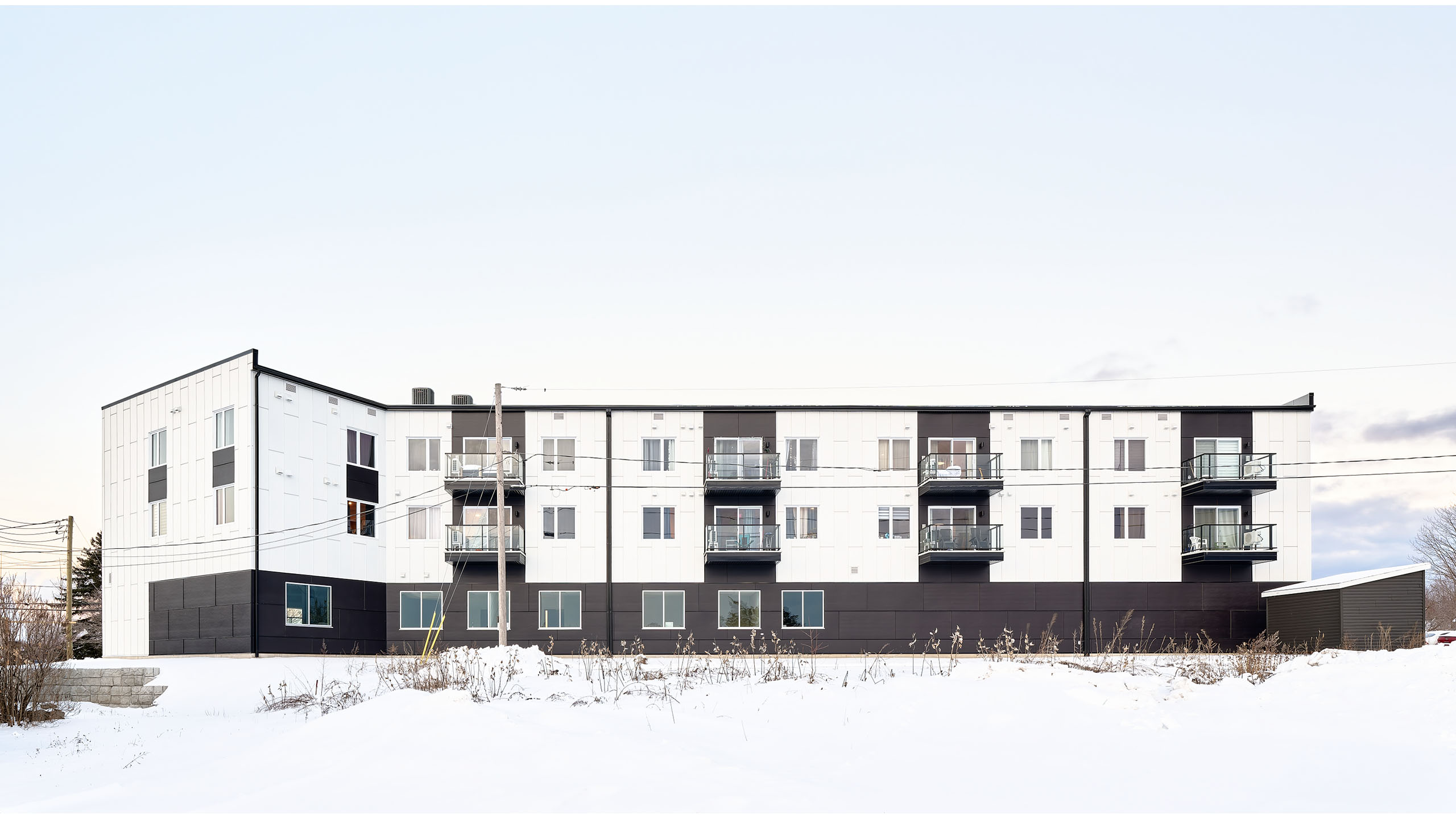 Un immeuble moderne de trois étages avec une façade blanche et noire se dresse dans un paysage enneigé et aride. Construction d'un nouveau bâtiment avec plusieurs balcons et de grandes fenêtres. On peut voir des lignes électriques et quelques plantes éparses au premier plan. Le ciel est couvert.