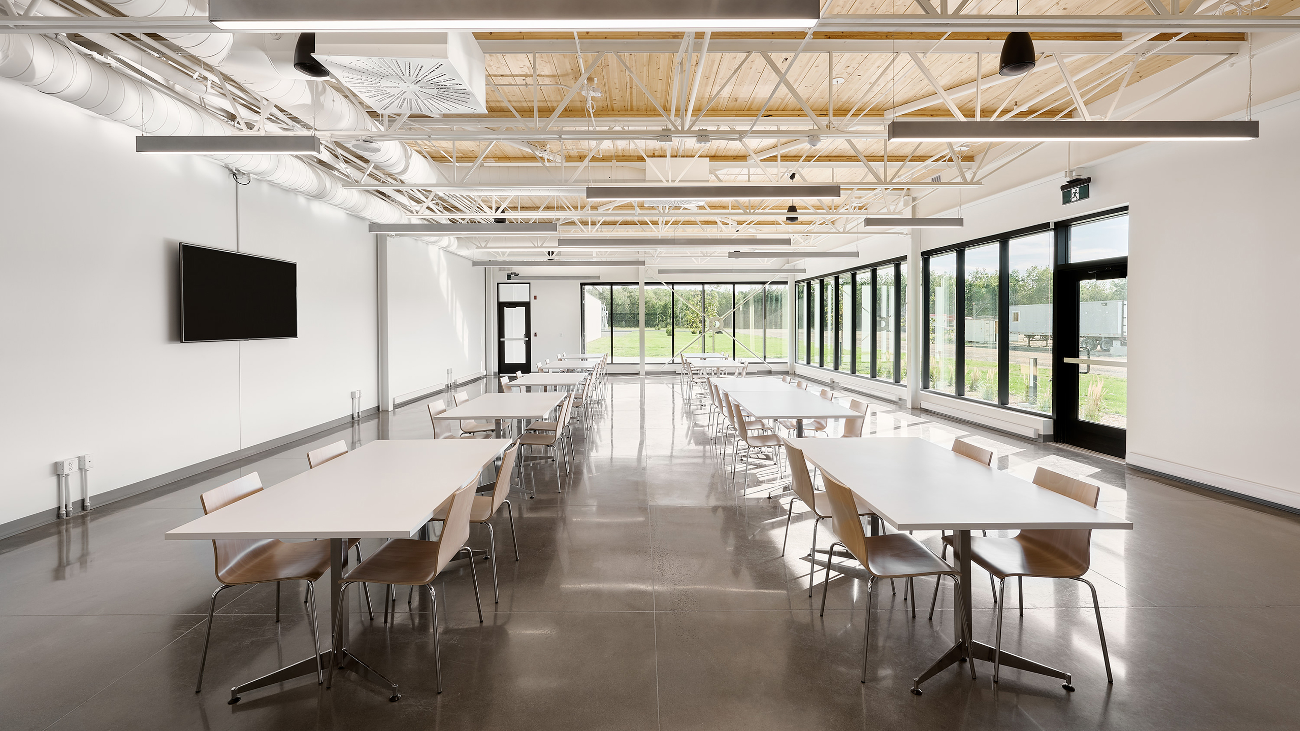 Une salle moderne et spacieuse avec de hauts plafonds et des poutres métalliques apparentes, rappelant la construction d'une nouvelle usine, dispose de plusieurs tables blanches et de chaises en bois. De grandes fenêtres sur un côté laissent entrer la lumière naturelle et une télévision à écran plat est fixée sur un mur blanc. Le sol est en béton poli.