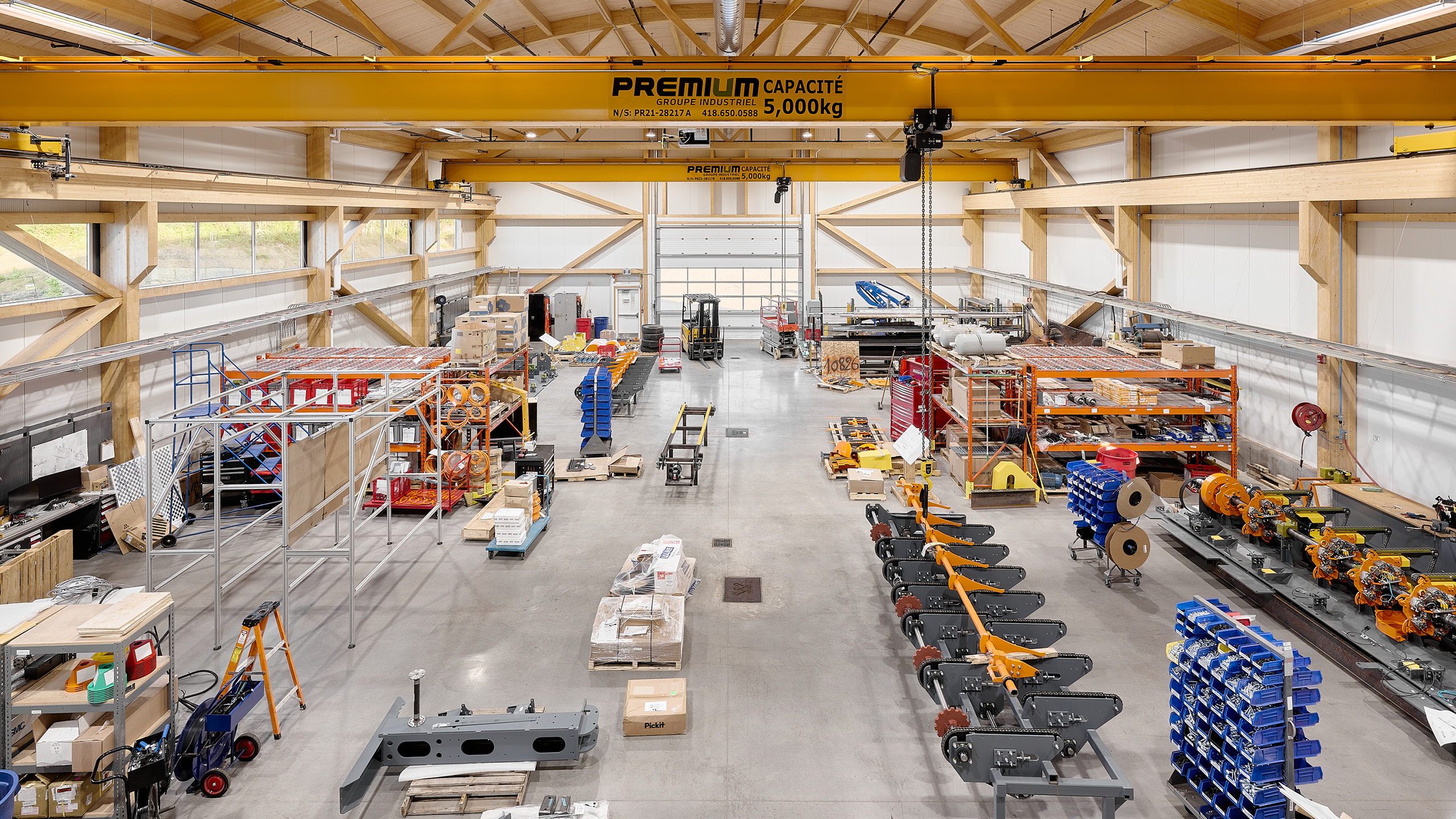 Un atelier industriel spacieux avec diverses machines, outils et matériaux disposés en sections. Au-dessus, une grande grue jaune étiquetée « PREMILUM CAPACITE 5 000 kg » traverse la pièce. L'espace est bien éclairé avec de grandes fenêtres et des poutres en bois soutenant le plafond, parfait pour la construction d'un nouveau siège social.