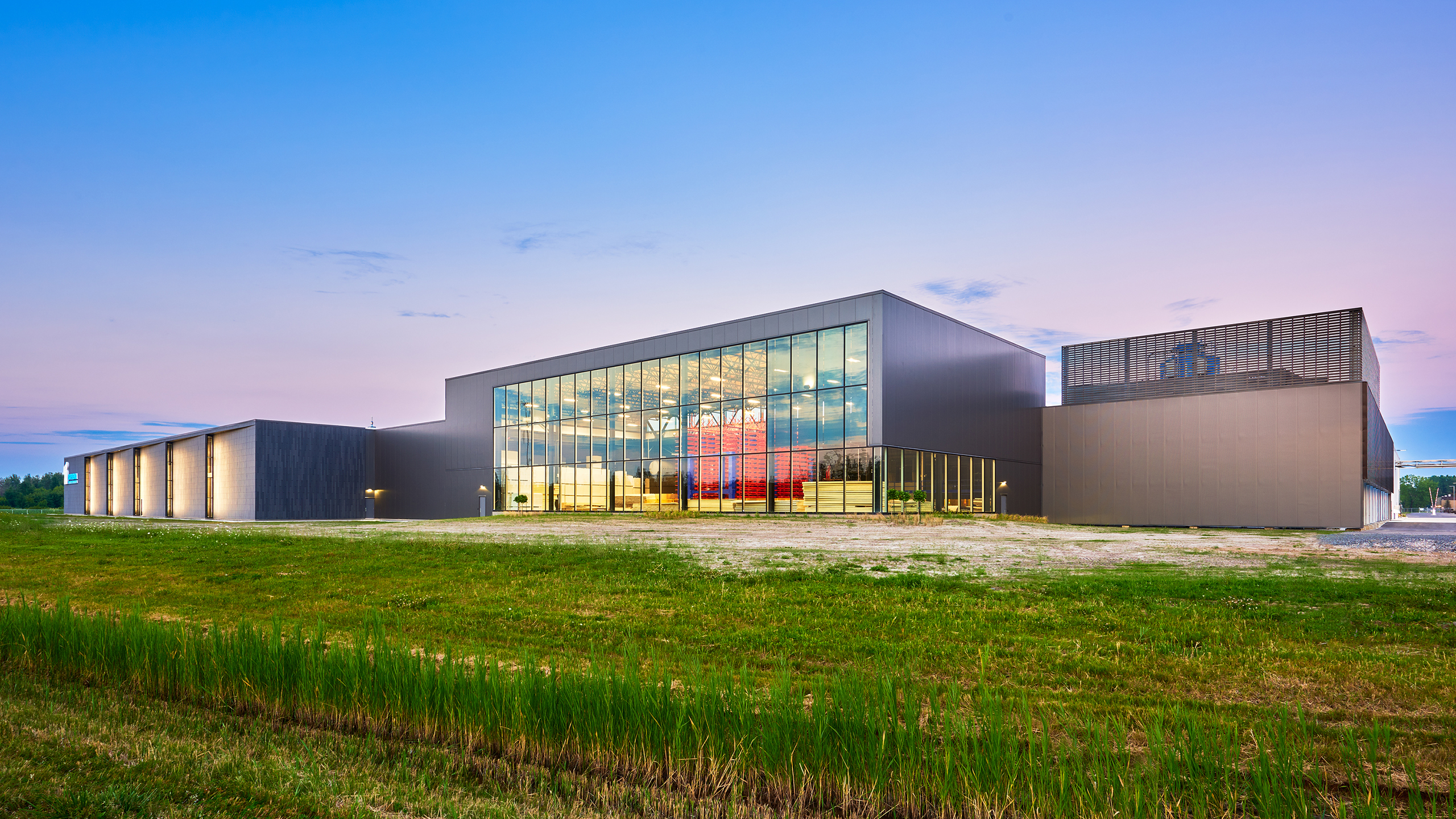 Un bâtiment moderne avec de grandes fenêtres en verre et des murs extérieurs sombres et épurés se dresse au milieu d'un vaste paysage verdoyant au crépuscule. Construction nouvelle usine se fond harmonieusement dans l'environnement serein. Le ciel est un doux dégradé de bleu et de rose, et le bâtiment est illuminé de l'intérieur, créant une lueur chaleureuse.