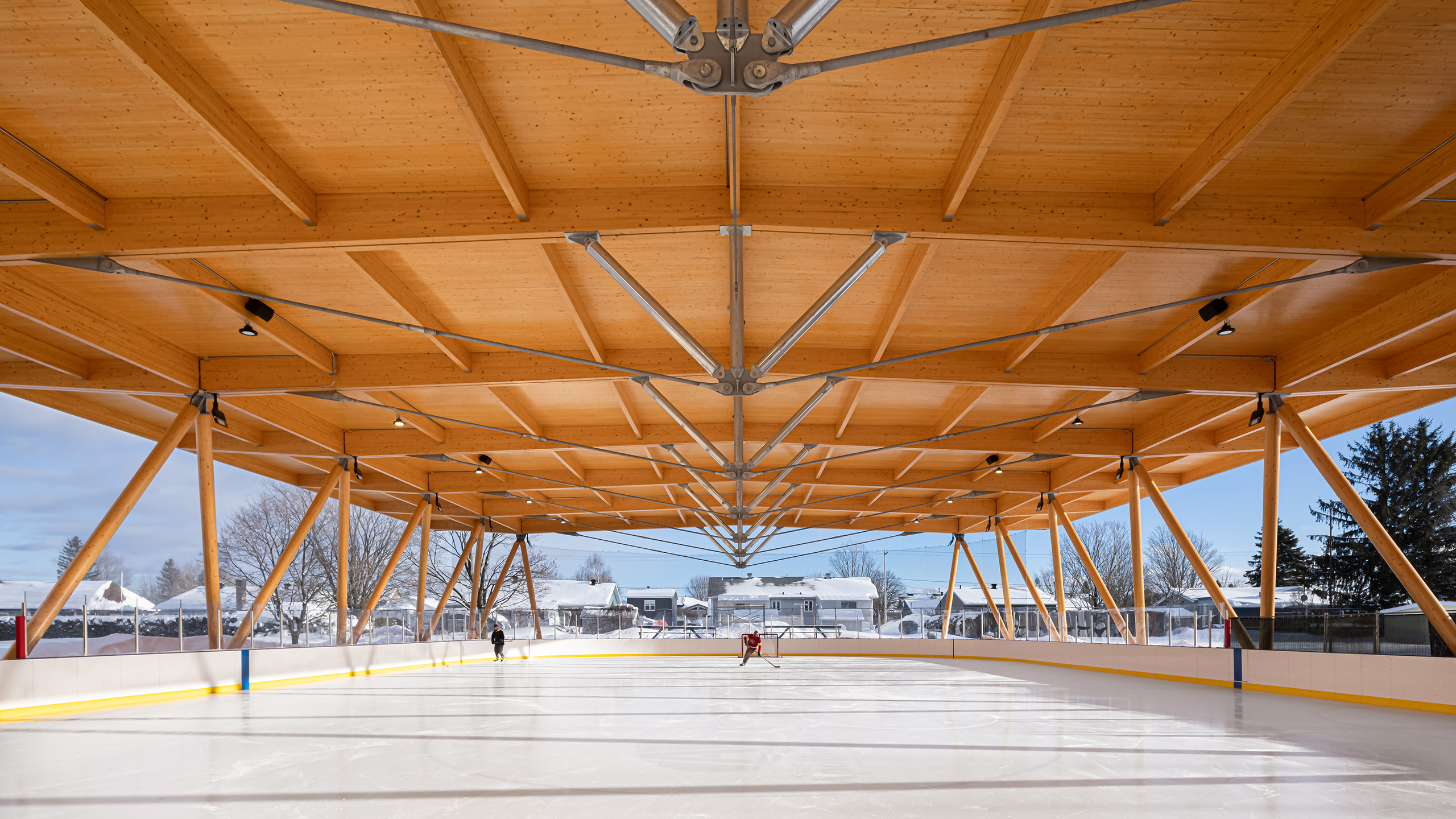 Une patinoire extérieure couverte, présentant une conception et une construction exquises, dispose d'un plafond à poutres en bois moderne avec de grandes fenêtres permettant à la lumière naturelle d'illuminer l'espace. Des arbres et des bâtiments recouverts de neige sont visibles en arrière-plan tandis que plusieurs personnes glissent sur la glace.