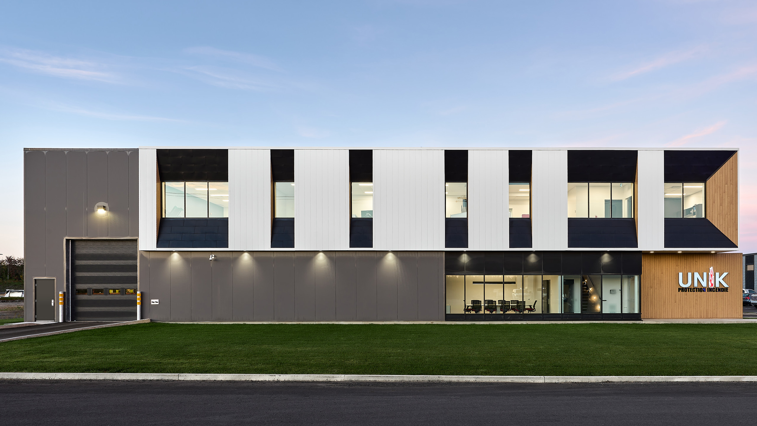 Bâtiment commercial moderne de deux étages avec de grandes fenêtres et un mélange de panneaux extérieurs gris, blancs et bois. Le niveau inférieur comprend un bureau aux parois vitrées et une grande porte de garage à gauche. La marque "UNIK" est visible sur le côté droit de la construction du nouveau siège social.