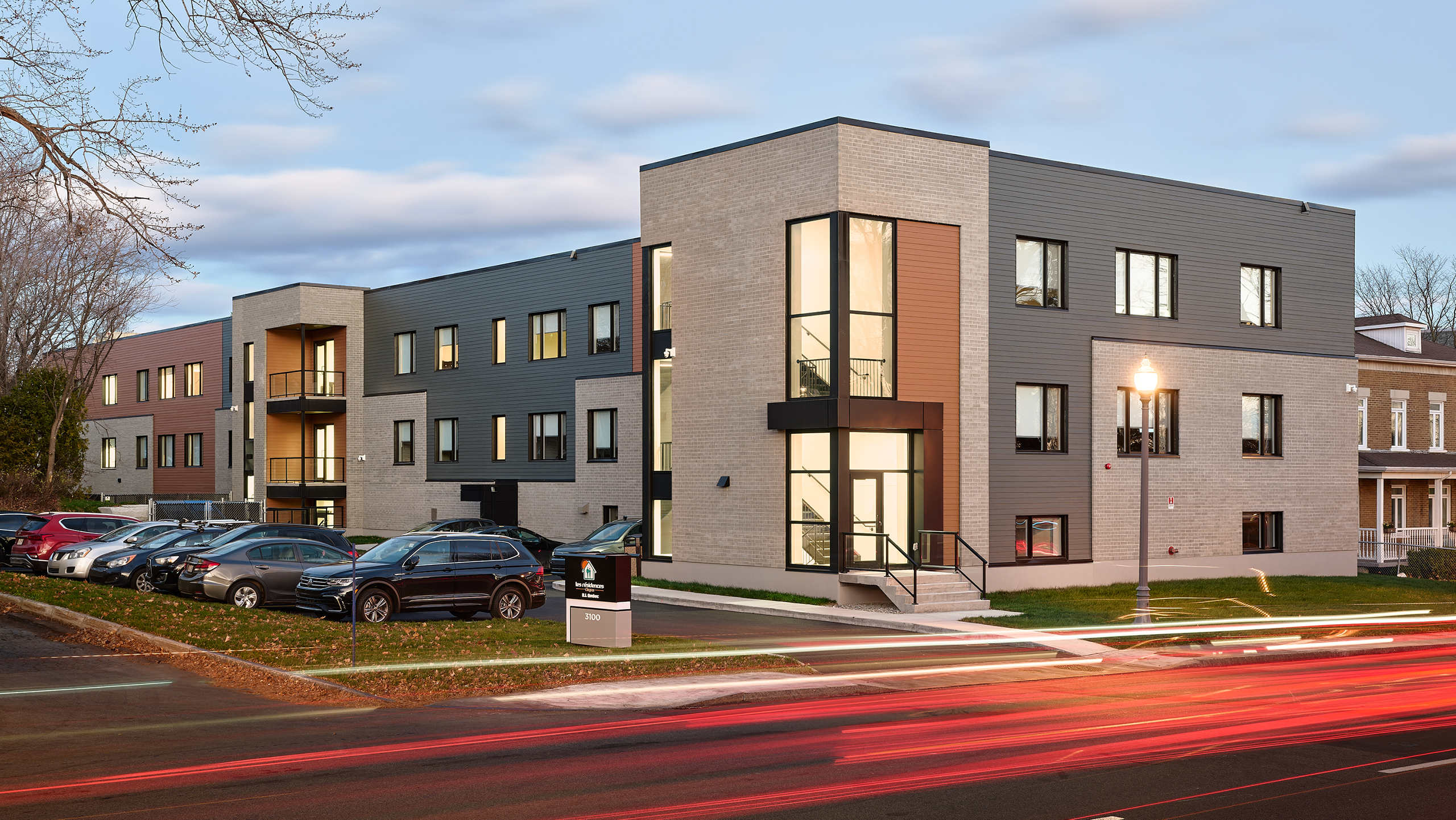 Immeuble d'appartements moderne de trois étages avec de grandes fenêtres et un mélange de panneaux extérieurs gris et beiges, mettant en valeur la construction d'un nouveau bâtiment. Une rangée de voitures garées borde la rue, qui est éclairée par des lampadaires. Les traînées lumineuses des véhicules qui passent créent des traînées dynamiques au premier plan.