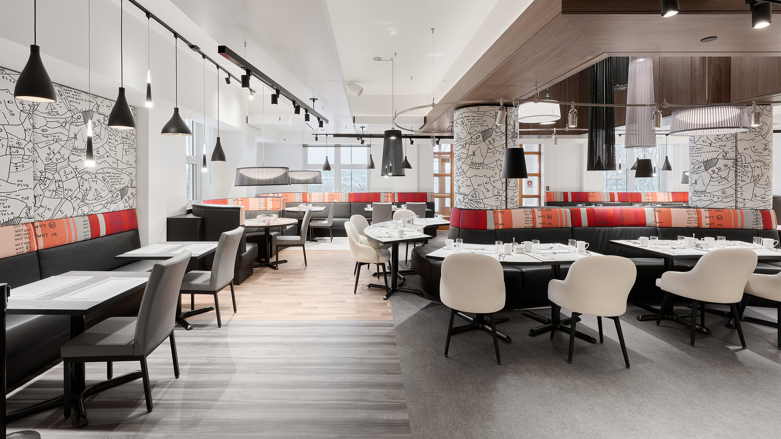 Un intérieur de restaurant moderne présentant un mélange de banquettes noires rembourrées et de chaises blanches tapissées autour de tables rondes et rectangulaires. Des luminaires suspendus sont suspendus au plafond et des illustrations artistiques en noir et blanc ornent les murs et les colonnes, lui donnant une touche sophistiquée similaire à celles que l'on trouve dans les résidences pour personnes âgées.