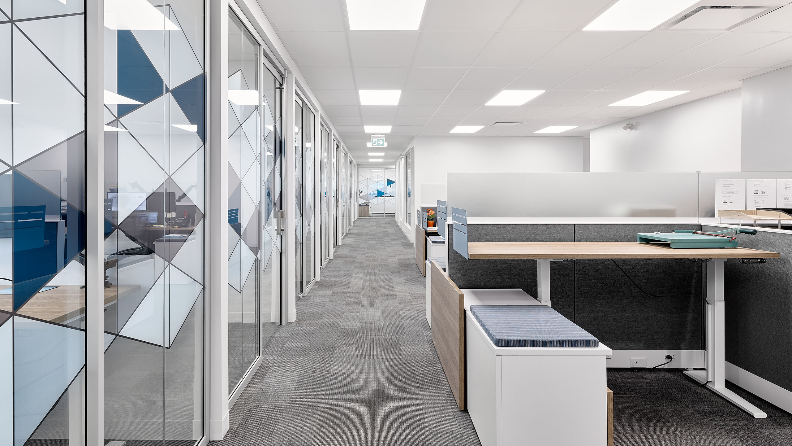 Un couloir de bureau moderne avec moquette au sol. À gauche, des bureaux privés avec des portes vitrées et des motifs géométriques. À droite, des cabines avec des bureaux debout et des fournitures de bureau. L'espace est bien éclairé par des plafonniers et l'aménagement général des bureaux est élégant et contemporain.