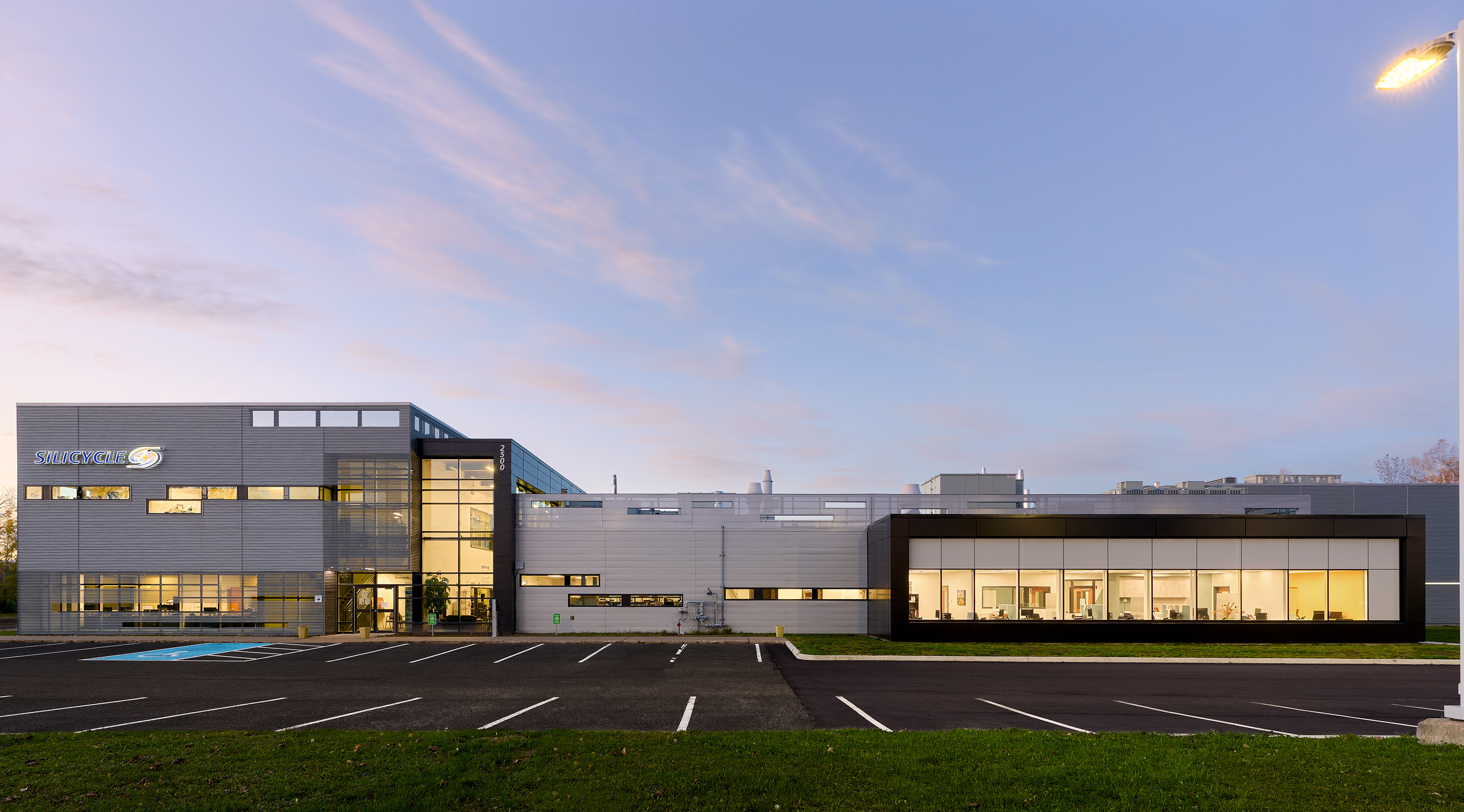 Vue grand angle d'un bâtiment industriel moderne avec de grandes fenêtres et un mélange de panneaux gris et beiges. Cette superbe construction industrielle est éclairée de l'intérieur, sous un ciel clair avec des reflets de coucher de soleil rosé. Un parking vide est au premier plan.