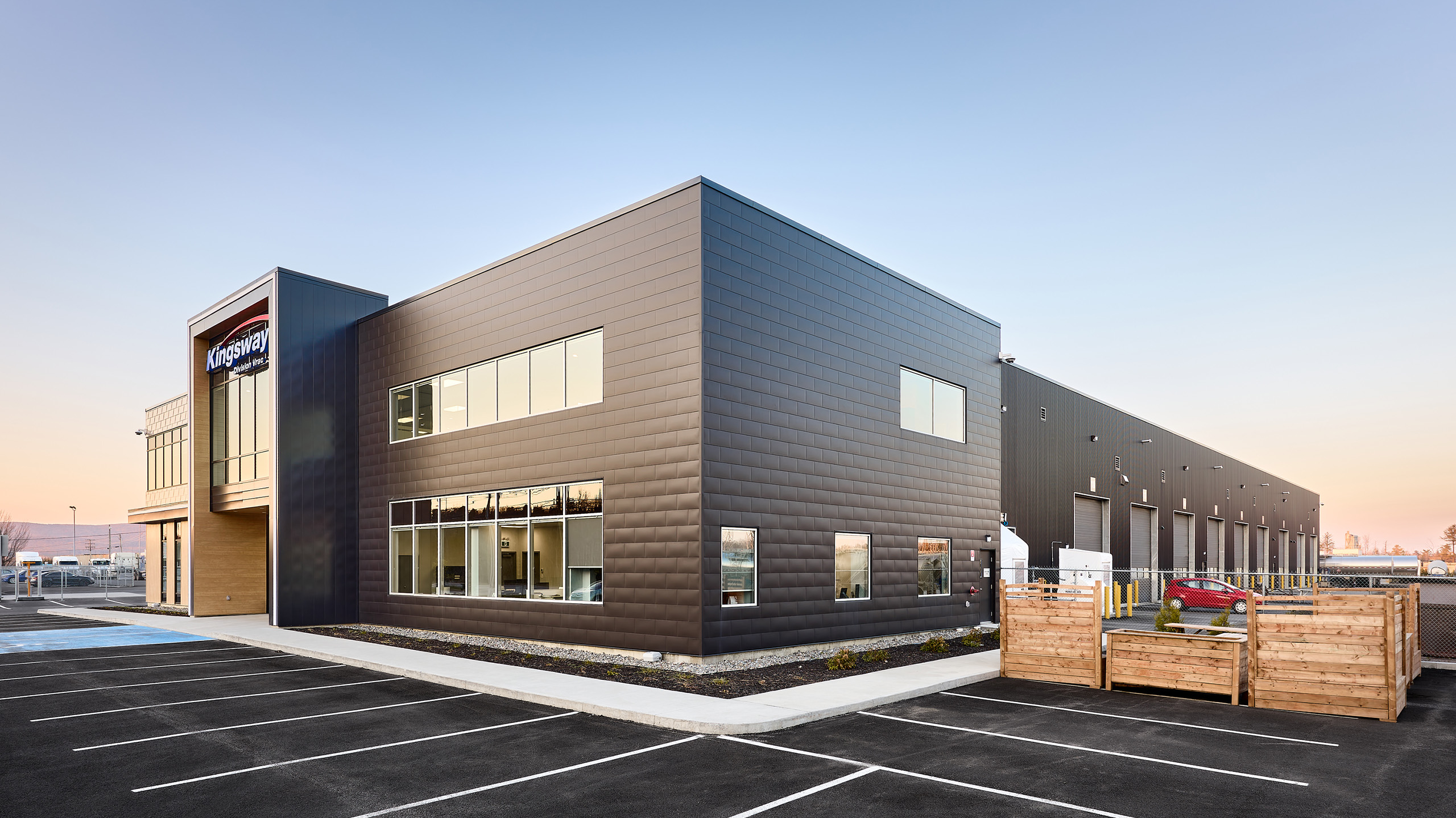 Un bâtiment industriel moderne avec un extérieur sombre et élégant et de grandes fenêtres à l'avant et sur les côtés. La construction d'un nouveau garage comprend plusieurs baies et un parking pavé spacieux sans véhicules. Le ciel est clair et bleu clair, suggérant le petit matin ou le soir.