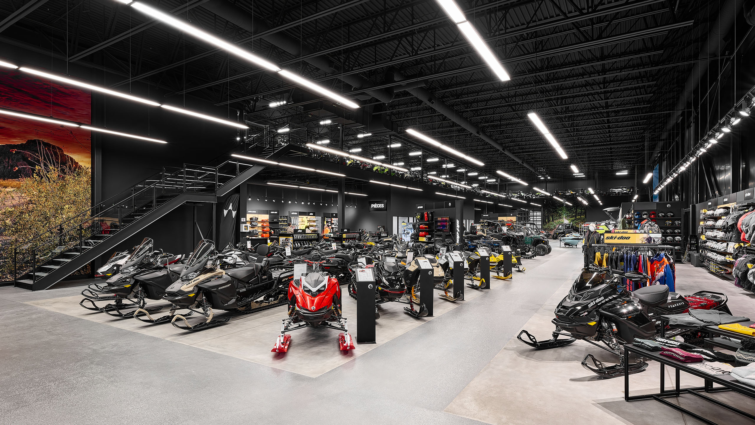 Une salle d'exposition élégante et moderne dans un bâtiment neuf présentant diverses motoneiges sous un éclairage vif. Les murs présentent des affiches et des équipements aux couleurs vives tandis que les étagères sont remplies d'accessoires et de vêtements. Un escalier mène à un niveau supérieur et le sol en béton poli renforce l'esthétique épurée.