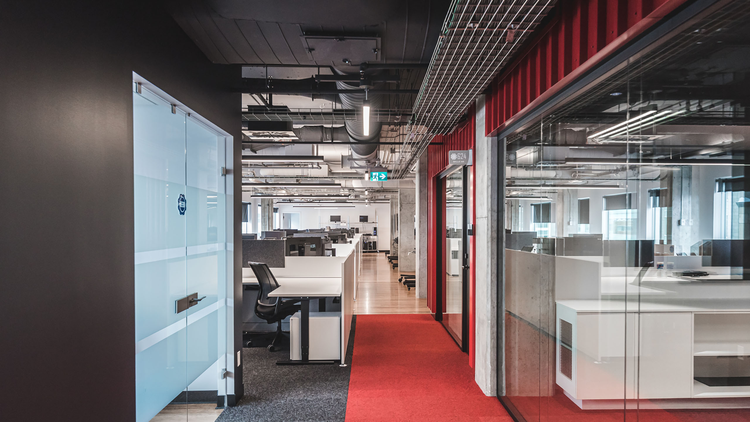 Un couloir de bureaux moderne, qui a récemment bénéficié d'une rénovation des espaces bureaux, présente une salle de réunion aux parois vitrées à droite et un espace de bureau ouvert rempli de bureaux et de chaises à l'arrière-plan. Le sol présente différentes textures, noir près de l'entrée et tapis rouge plus loin. L'espace est bien éclairé.