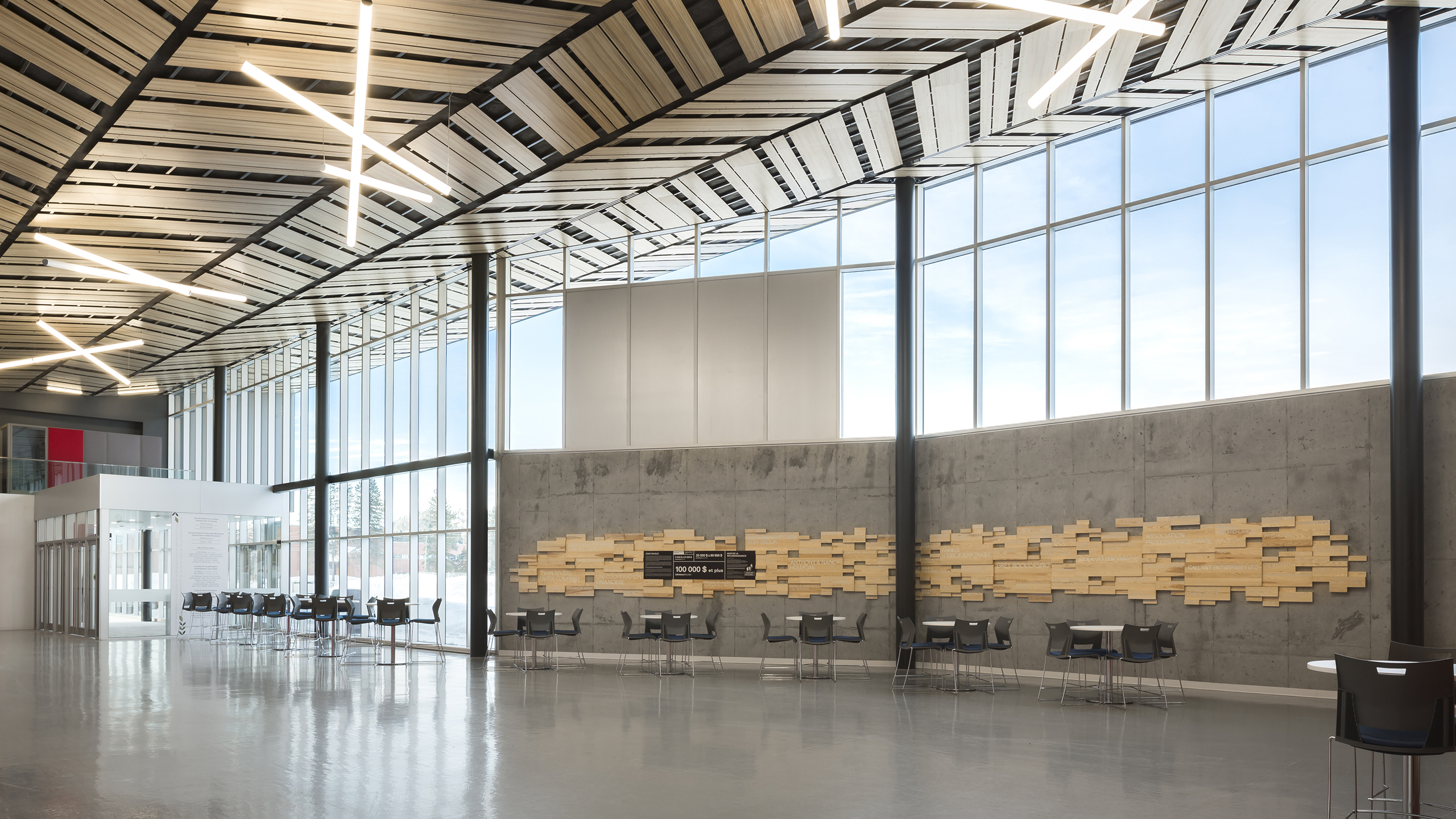 Un hall d'entrée moderne et spacieux avec de hauts plafonds, de grandes fenêtres et des chaises et tables dispersées le long des murs ressemble à un amphithéâtre serein. Le plafond est orné de boiseries uniques et de luminaires contemporains. Un mur présente une installation artistique abstraite en bois.